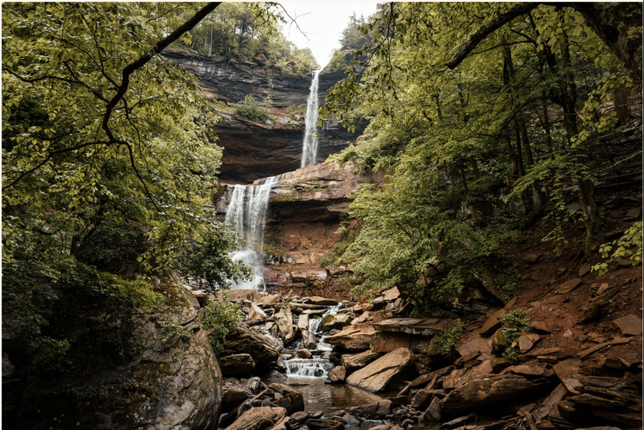 Waterfall cascading down a rocky cliff in a lush, green forest with a stream flowing over rocks at the base.