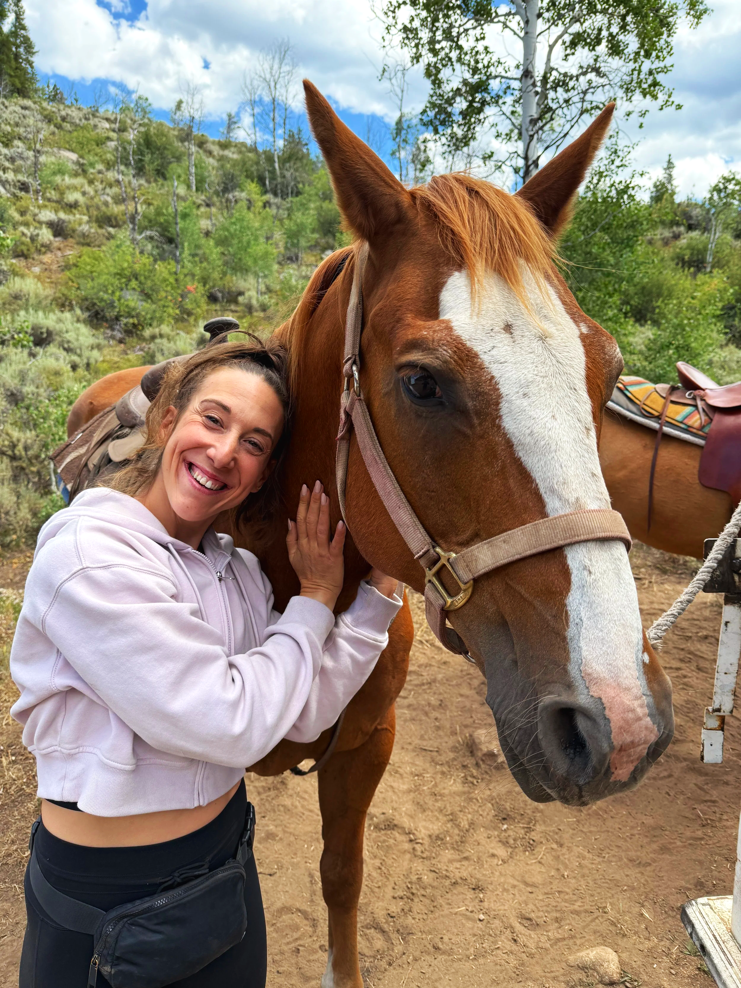 A woman smiling and hugging a brown horse with a white face marking outdoors in a wooded area with mountains and trees in the background.