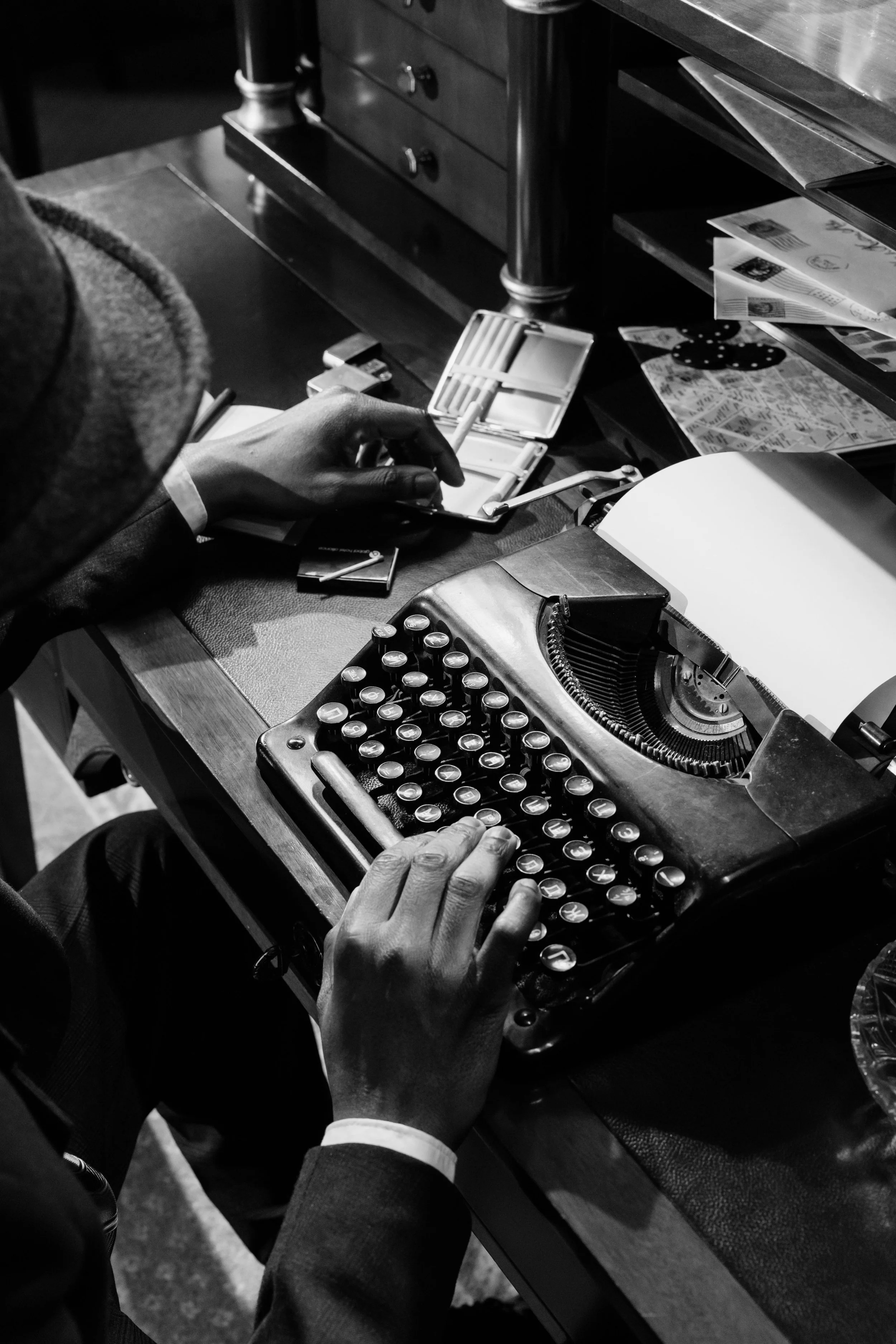 A person in a suit using a vintage typewriter on a desk with papers and office supplies in black and white.