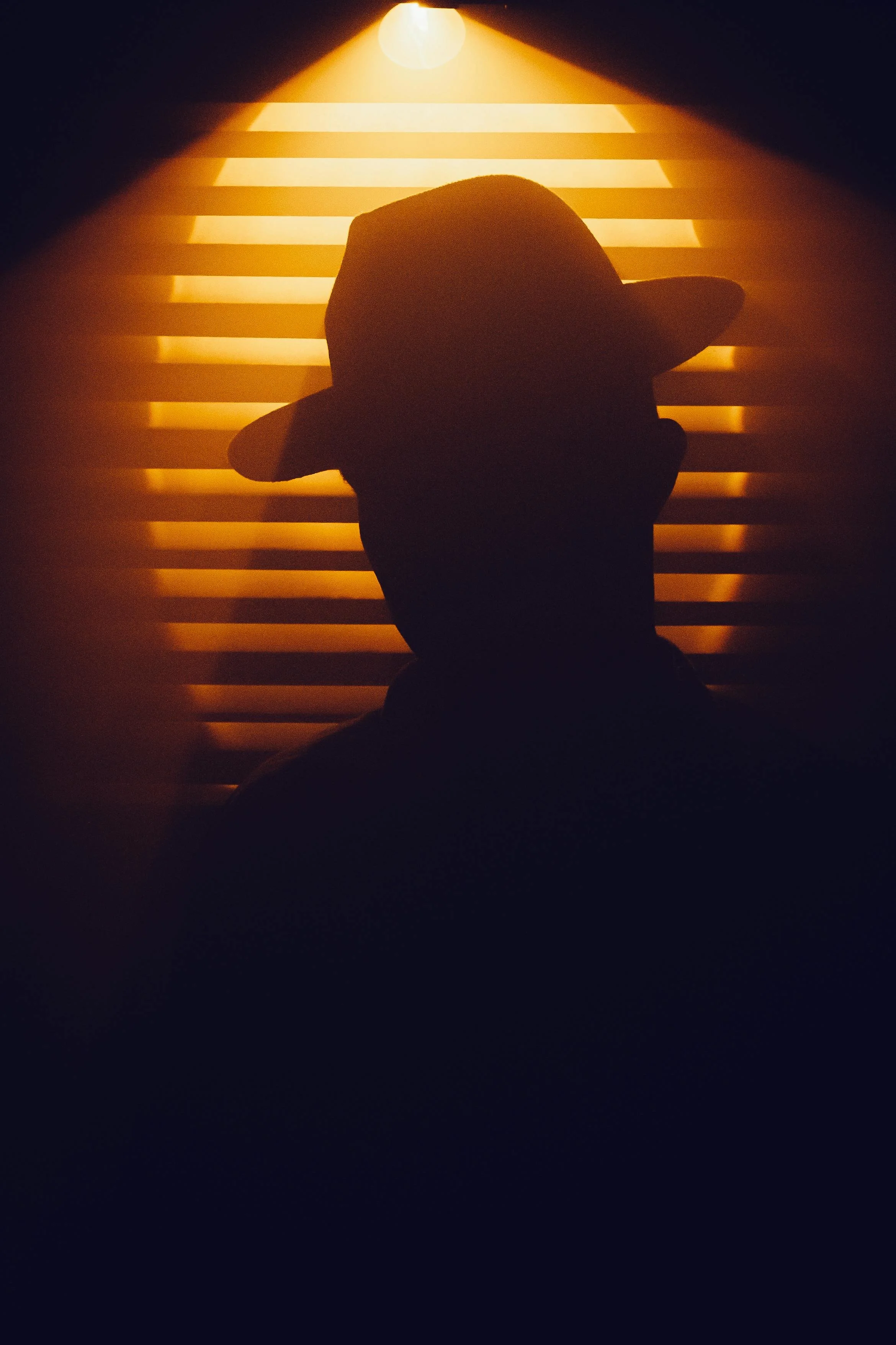 Silhouette of a person wearing a hat, standing in front of window blinds with sunlight shining through.