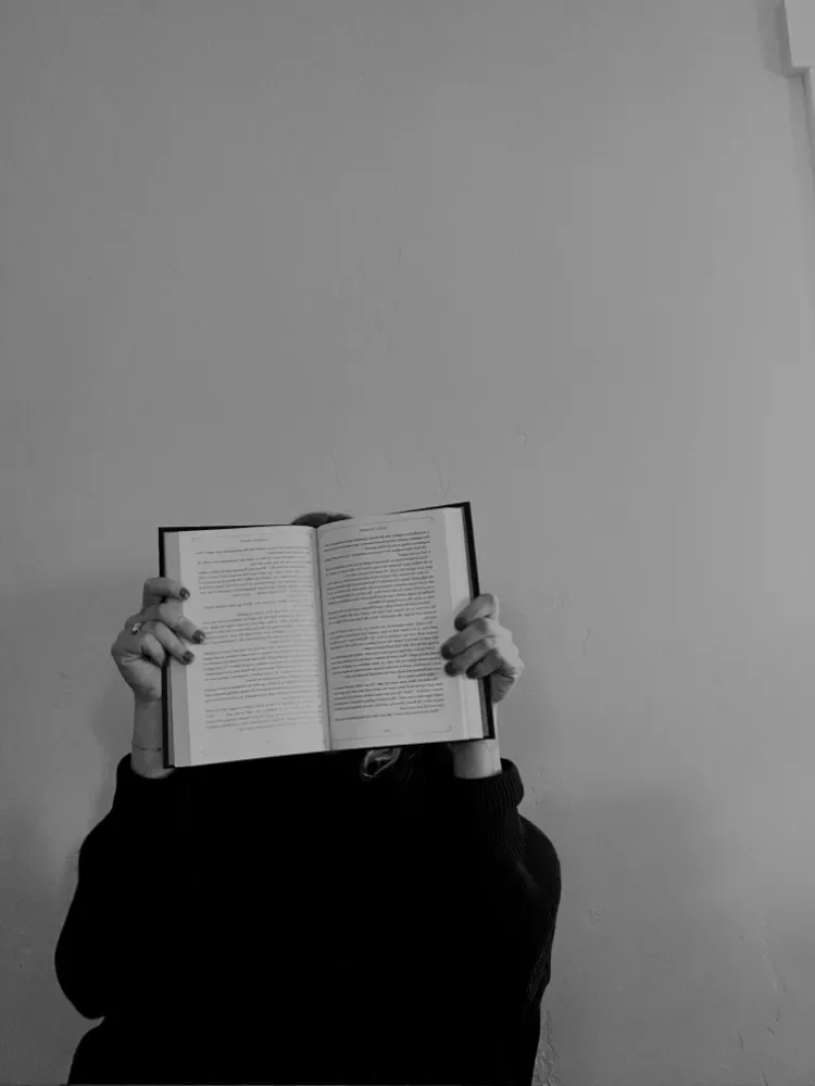 Person holding an open book in front of their face in front of a plain wall