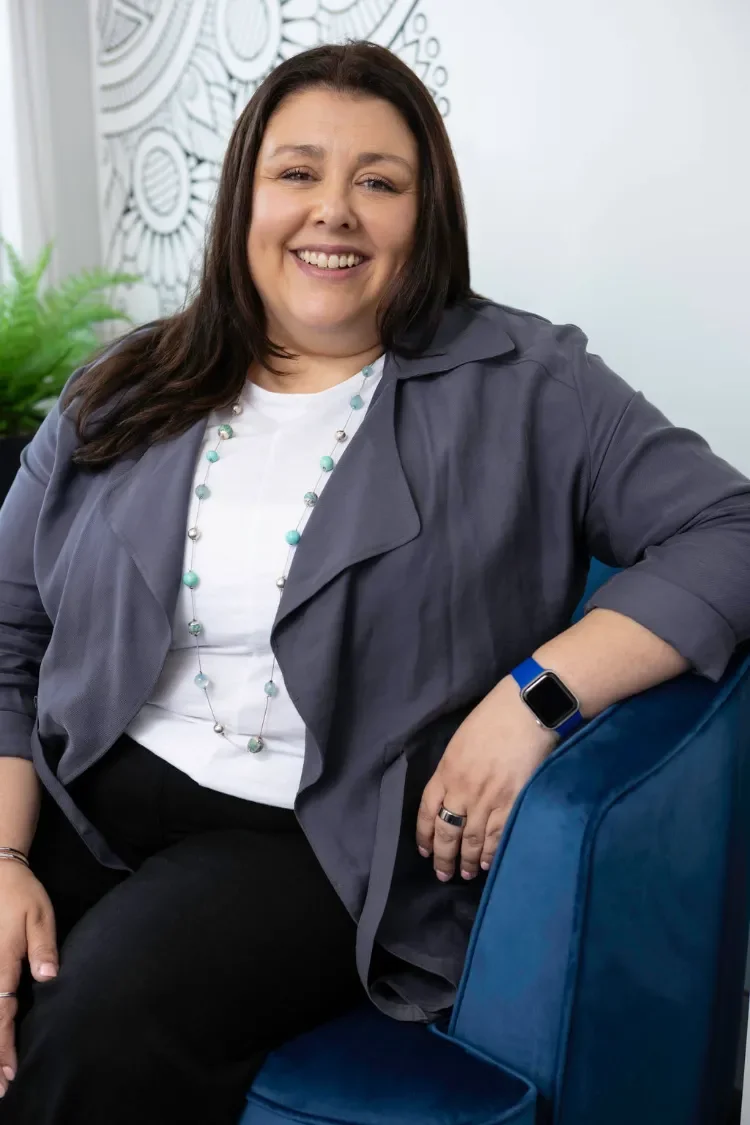 Three Pillars Training centre director, Sharon Williams with long dark hair, wearing a white top, a blue-grey jacket, and beaded necklace sitting on a blue chair.