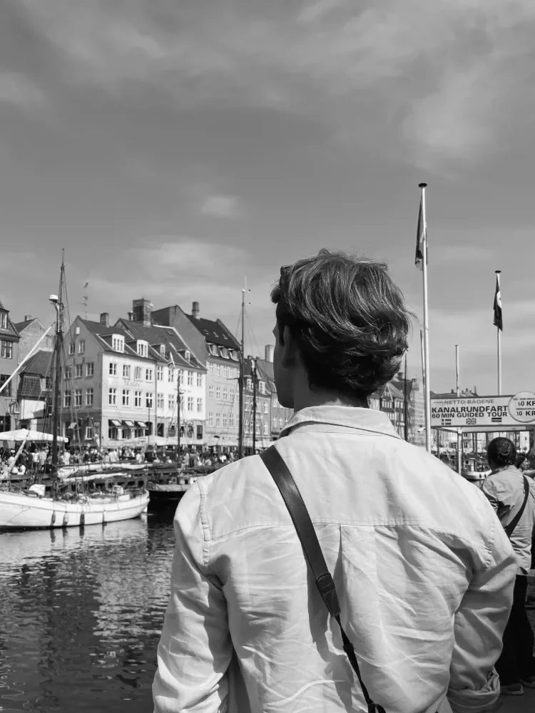 A man with short hair and a white jacket stands near a harbour, looking at the boats and historic buildings, with flags and a cloudy sky in the background.