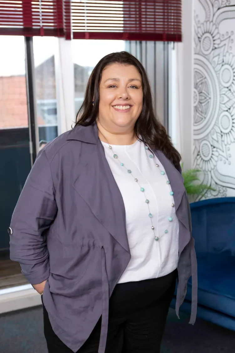 Three Pillars Training Director with long dark hair, wearing a blue-grey jacket over a white shirt, standing in the Lincoln training centre near a window with blinds and a patterned wall in the background.