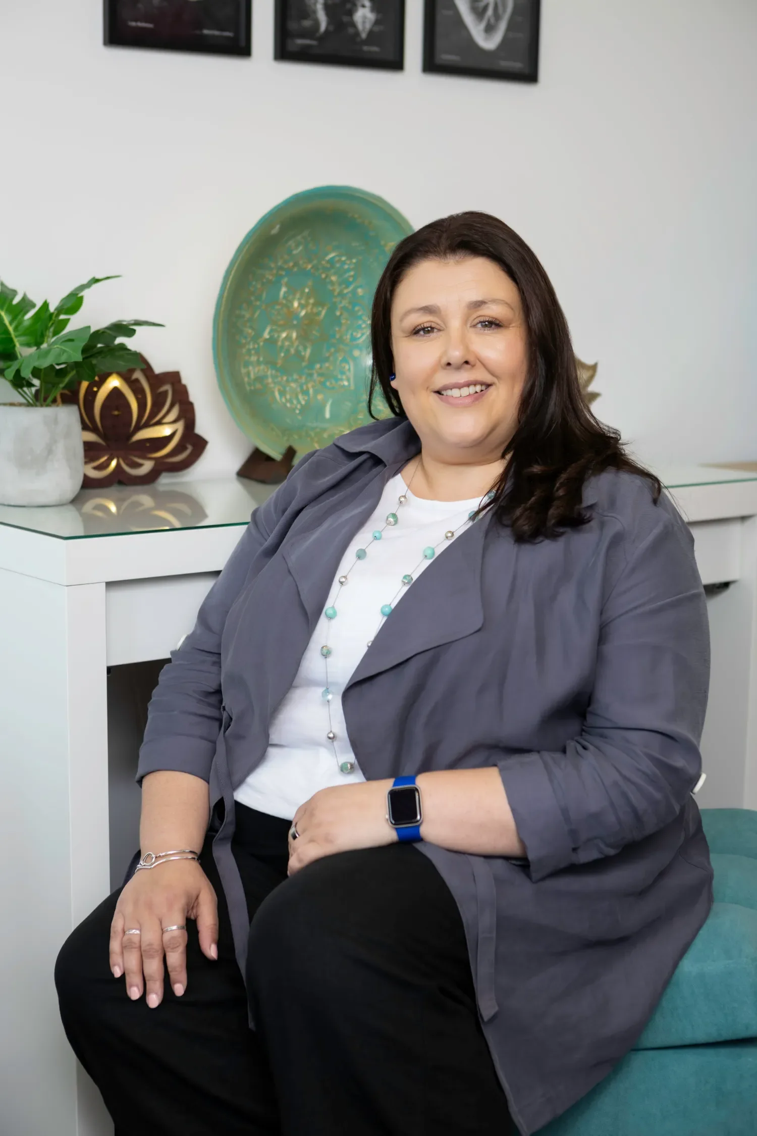 A woman with long dark hair, wearing a gray jacket, white shirt, and turquoise necklace, sitting on a teal chair in a modern office or living space. In the background, there are decorative items including a plant, a green tray, and framed artwork on the wall.