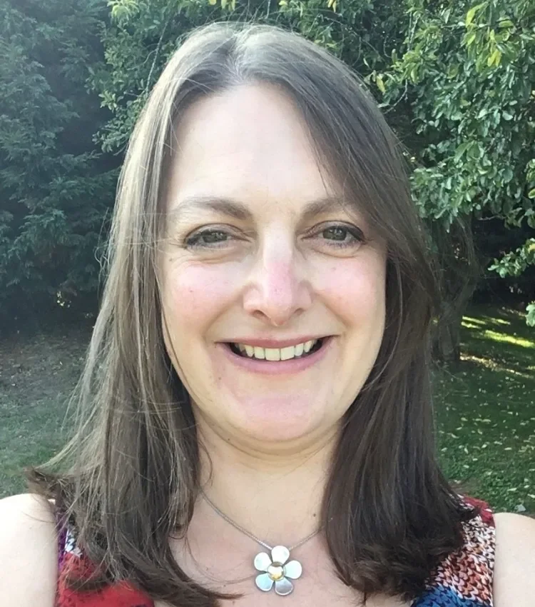A woman with shoulder-length brown hair smiling outdoors with green trees in the background, wearing a floral necklace and a red patterned top.