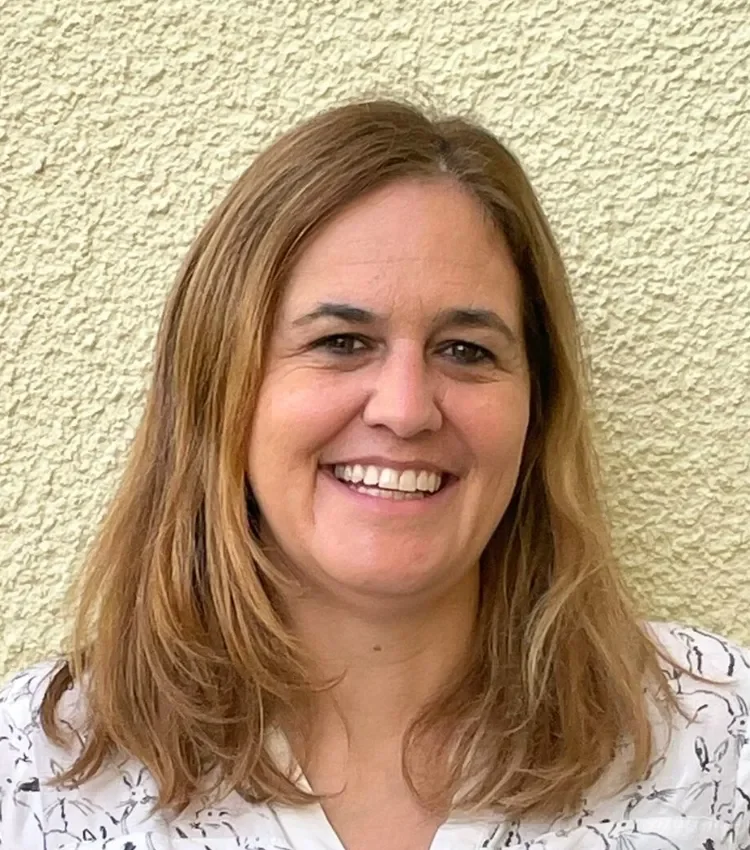 A woman with shoulder-length light brown hair smiling in front of a textured beige wall.