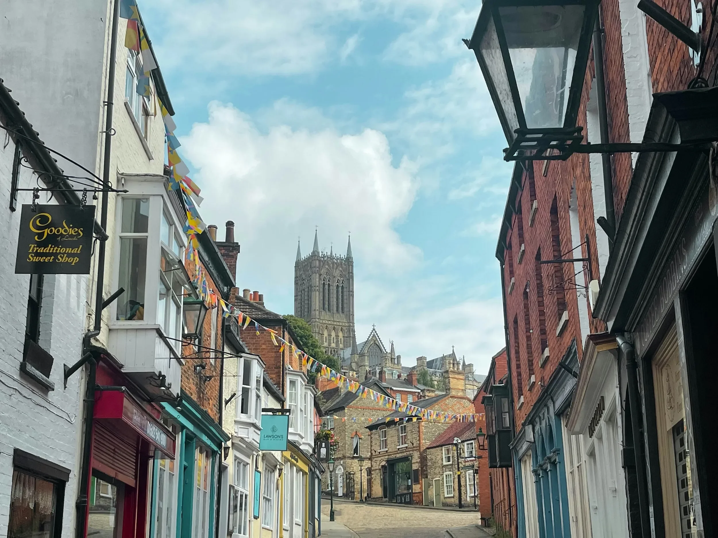 A narrow street in Lincoln city centre lined with shops, with colourful bunting hanging across, and Lincoln cathedral in the background under a partly cloudy sky.