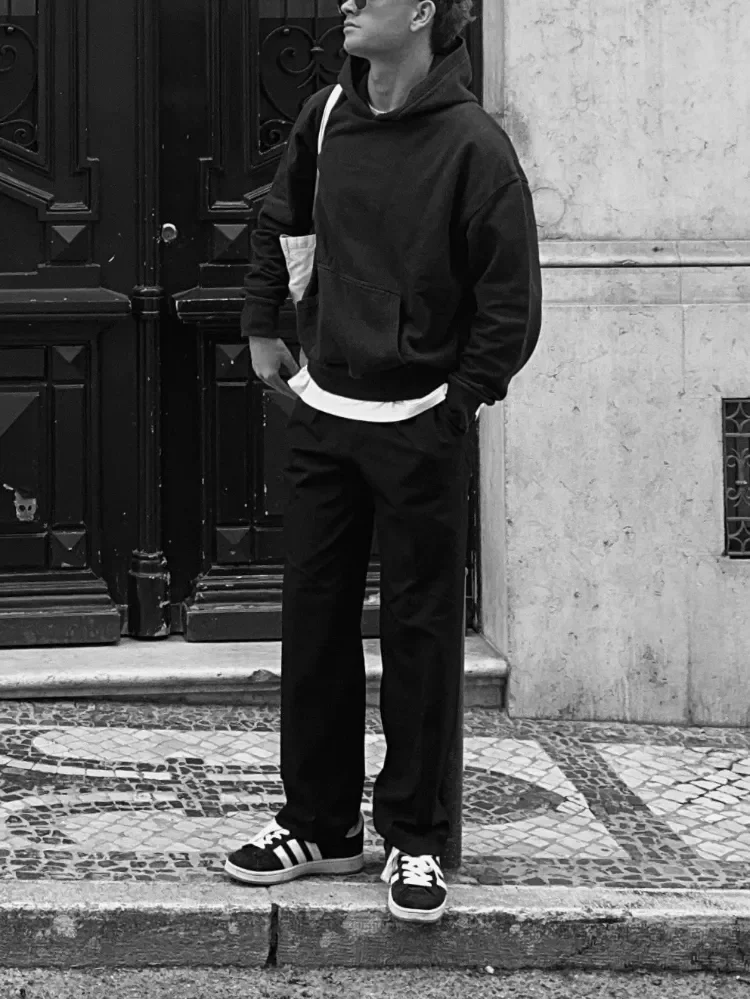 A black and white image of a man dressed in a hoodie and trainers standing on the side of a tiled pavement
