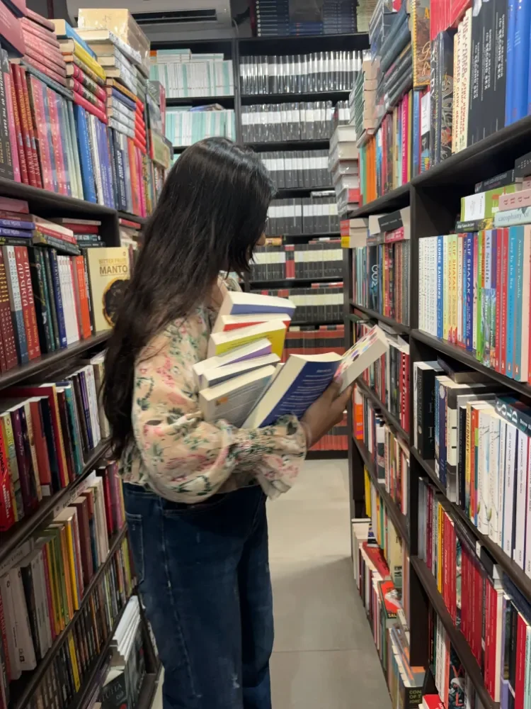 A woman with long dark hair in a library, holding several books and browsing through the shelves.