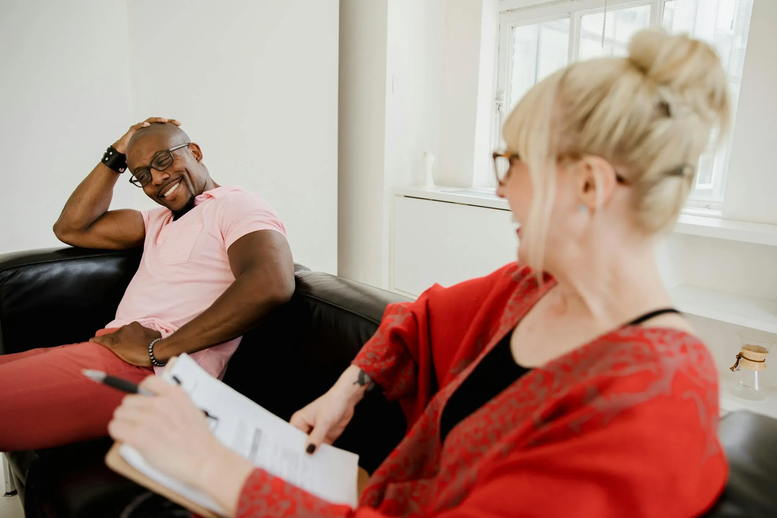 A man in a pink shirt is sitting on a black leather couch, smiling and touching his head, in conversation with a woman with blonde hair in a bun, glasses, and a red jacket, who is writing on a notepad.