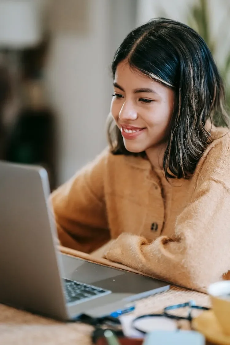 Young woman with dark hair smiling while using a laptop in a cozy indoor setting.