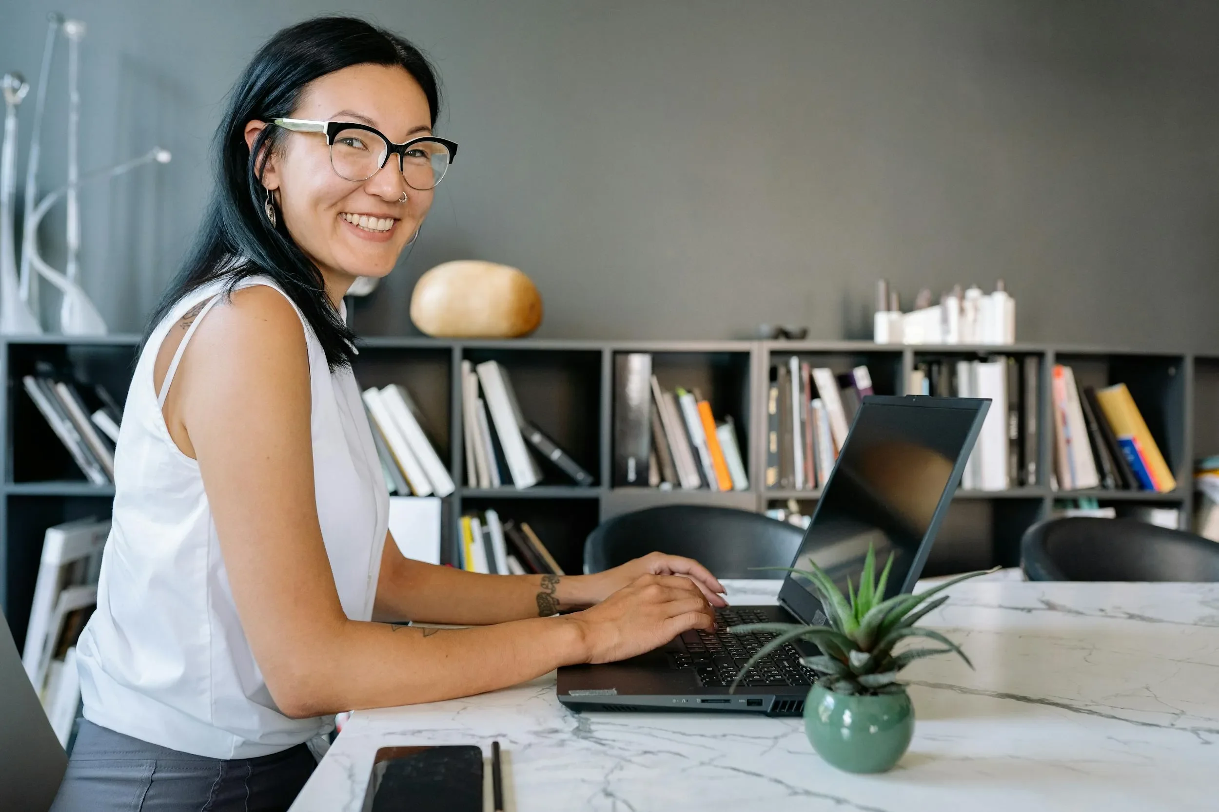 A woman with glasses and dark hair smiling while working on a laptop at a marble table with a potted plant with a bookcase in the background.