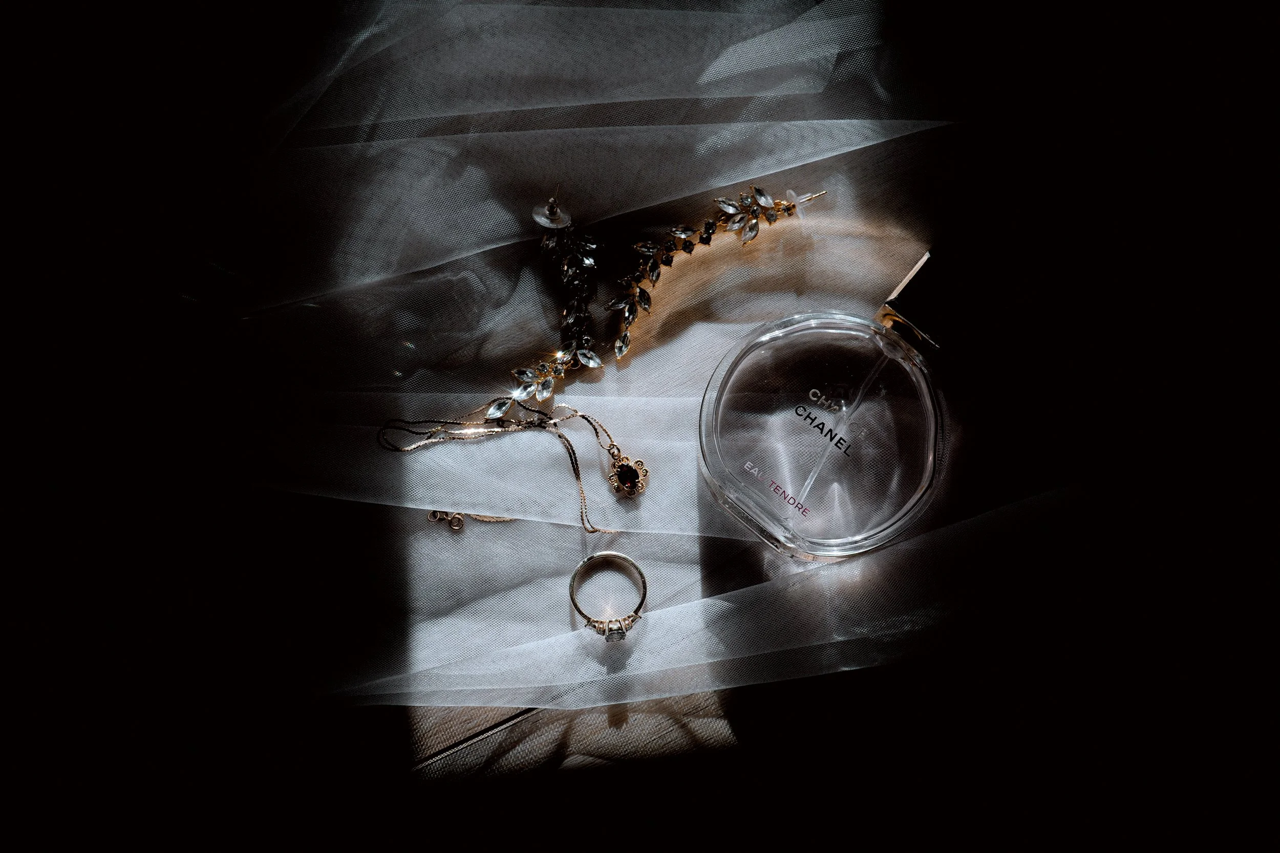 Jewelry, including a necklace, ring, and earrings, beside a partially pressed Chanel perfume bottle on a sheer fabric background with dramatic lighting.