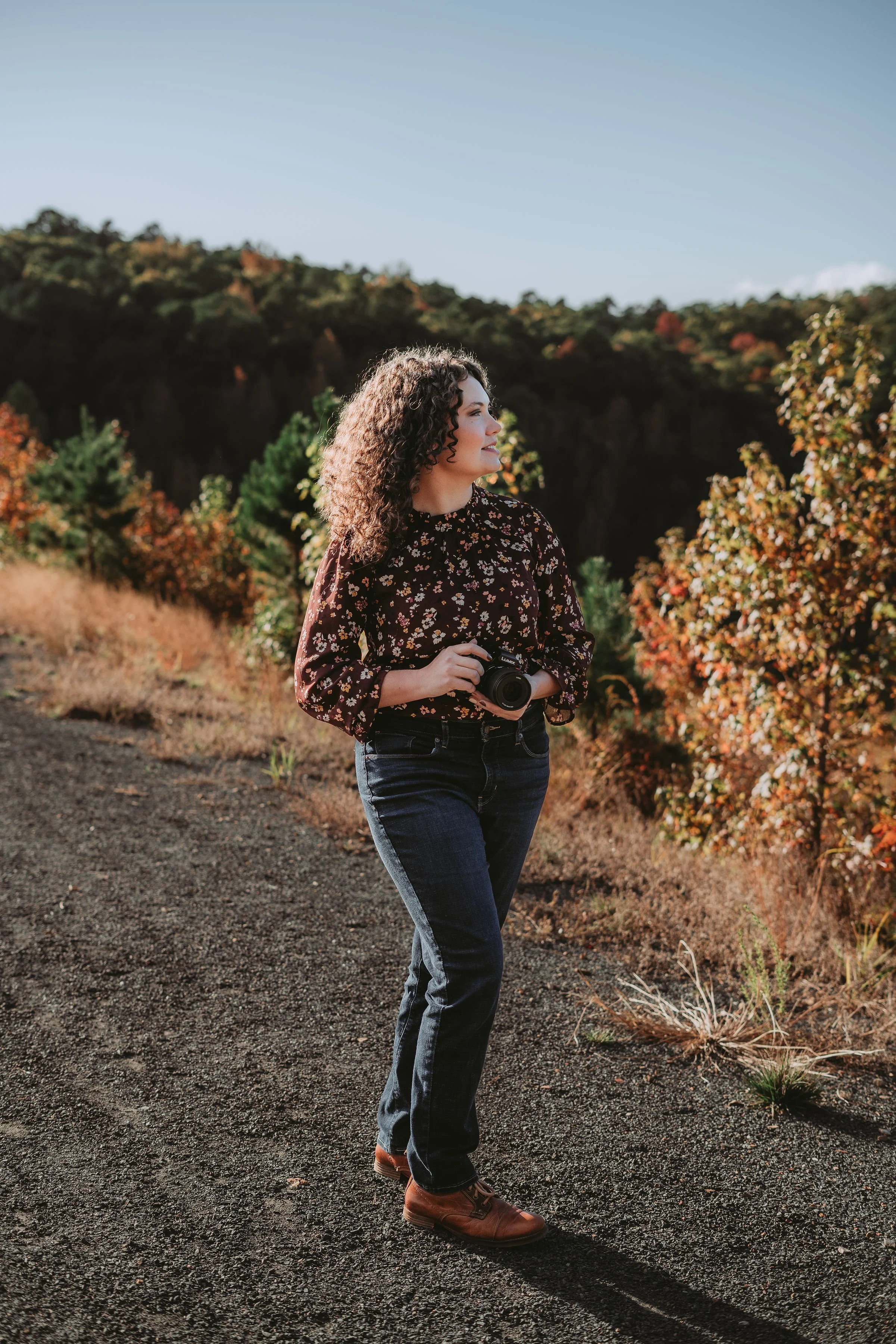 A woman with curly hair holding a camera, standing on a dirt path in a scenic outdoor area with trees and hills, during daytime.