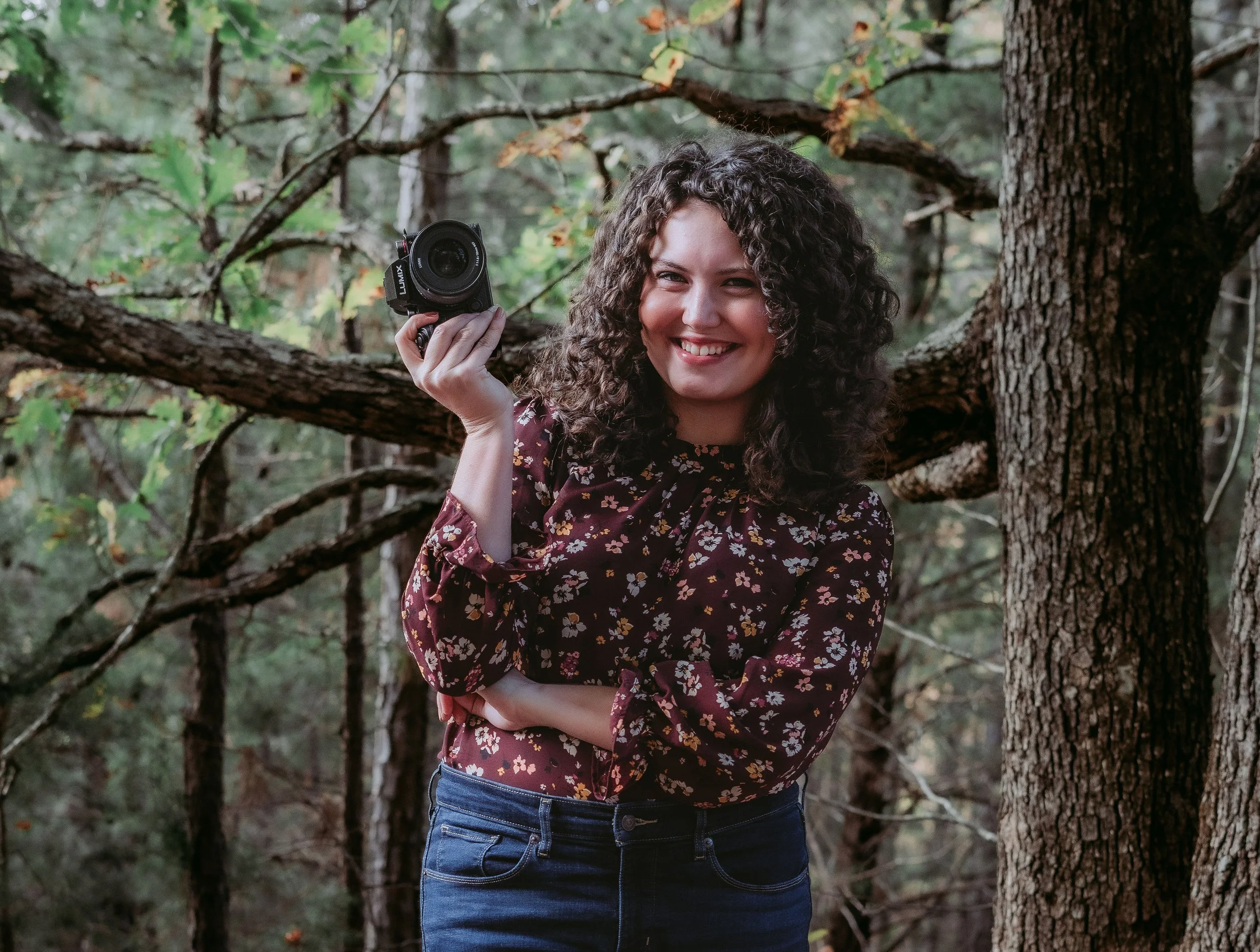 A woman with curly hair smiling and holding a camera in a forest.
