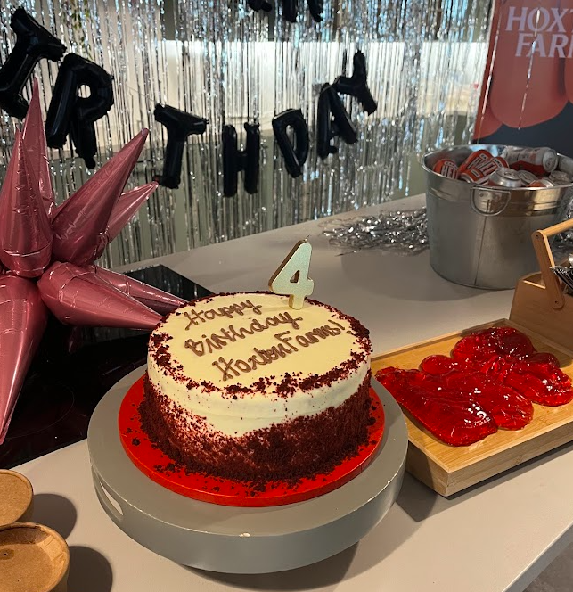 A birthday cake with a white and red velvet design, topped with a number 4 candle, on a white table. Behind it are balloons spelling 'HAPPY BIRTHDAY' and a silver balloon, with party supplies nearby.