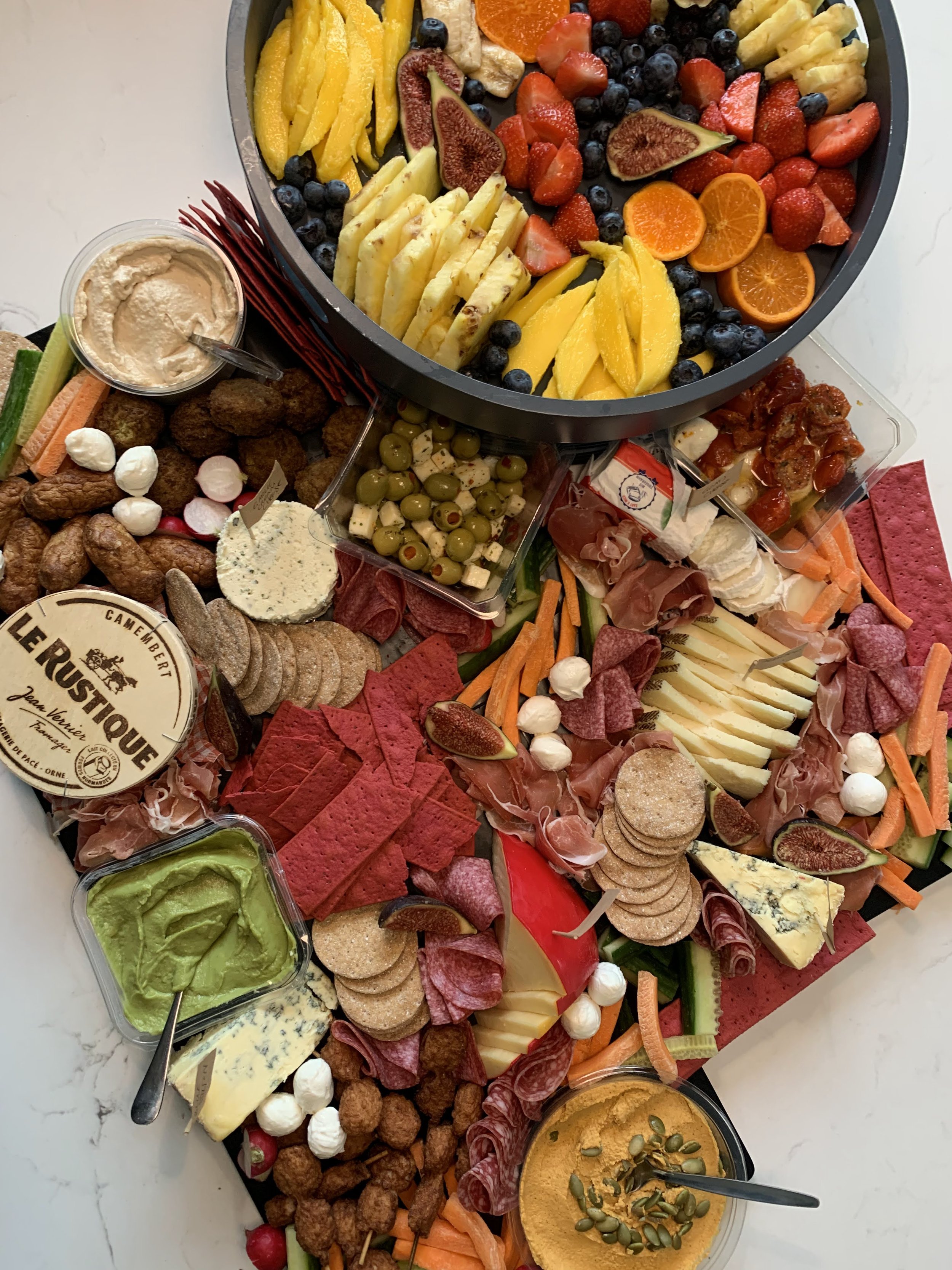 An assortment of fresh fruits, cheeses, charcuterie, dips, and snacks on a table, including sliced pineapple, strawberries, blueberries, figs, mandarin oranges, and various cheeses and meats, arranged on a platter.