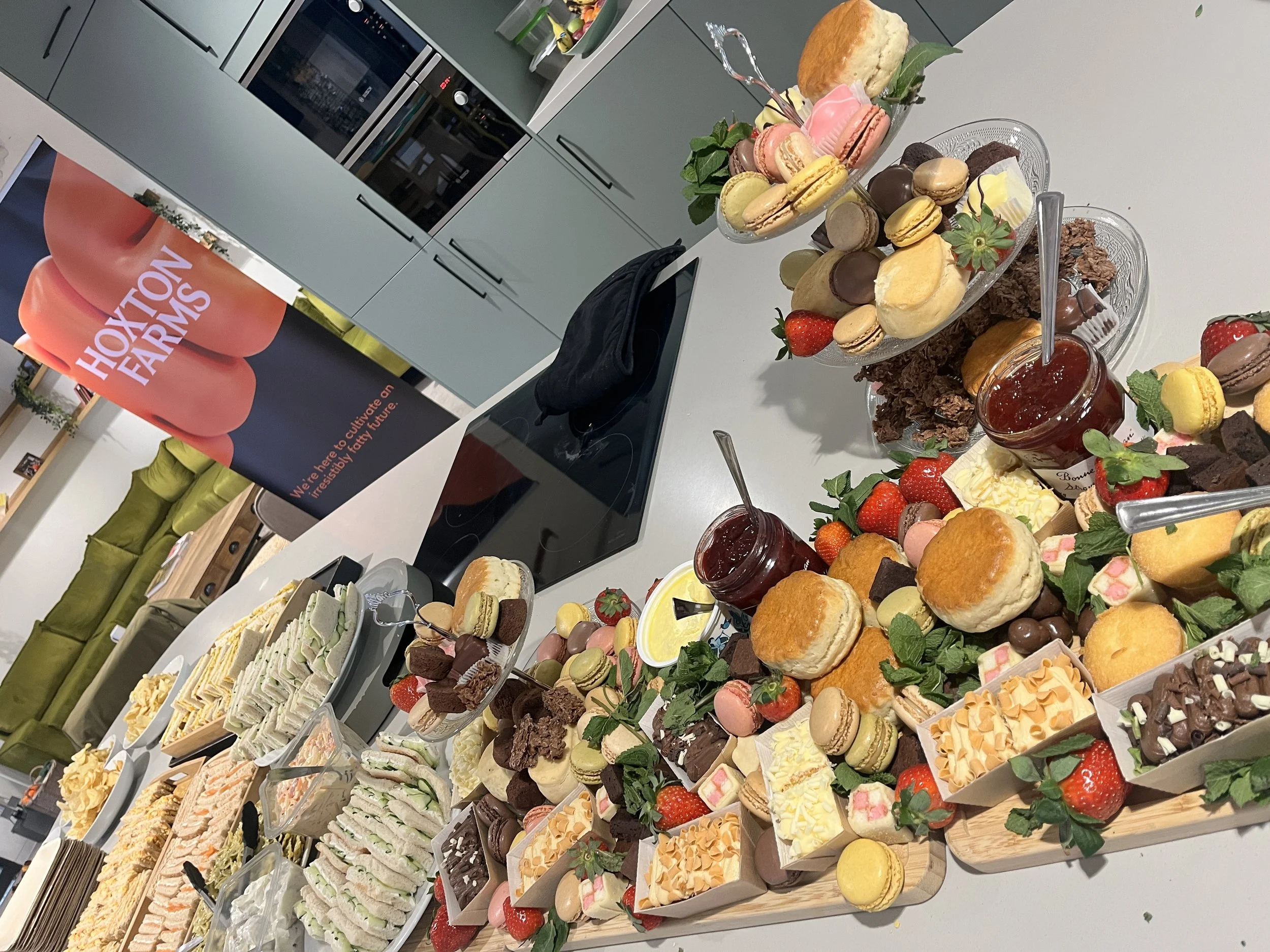 A spread of assorted desserts and finger foods on a kitchen counter, including mini cakes, macarons, strawberries, jam jars, and other sweets, with a Hoxiton Farms sign in the background.