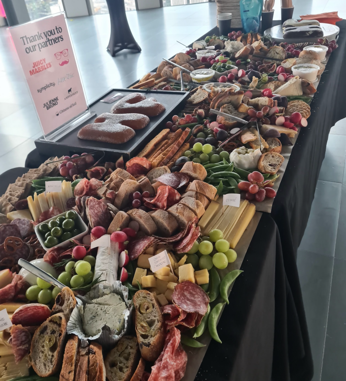 A long table with a black tablecloth displays an assortment of cheeses, cured meats, grapes, crackers, and dips, arranged for a buffet or tasting event. A sign on the table thanks partners such as Juicy Marbles and others.