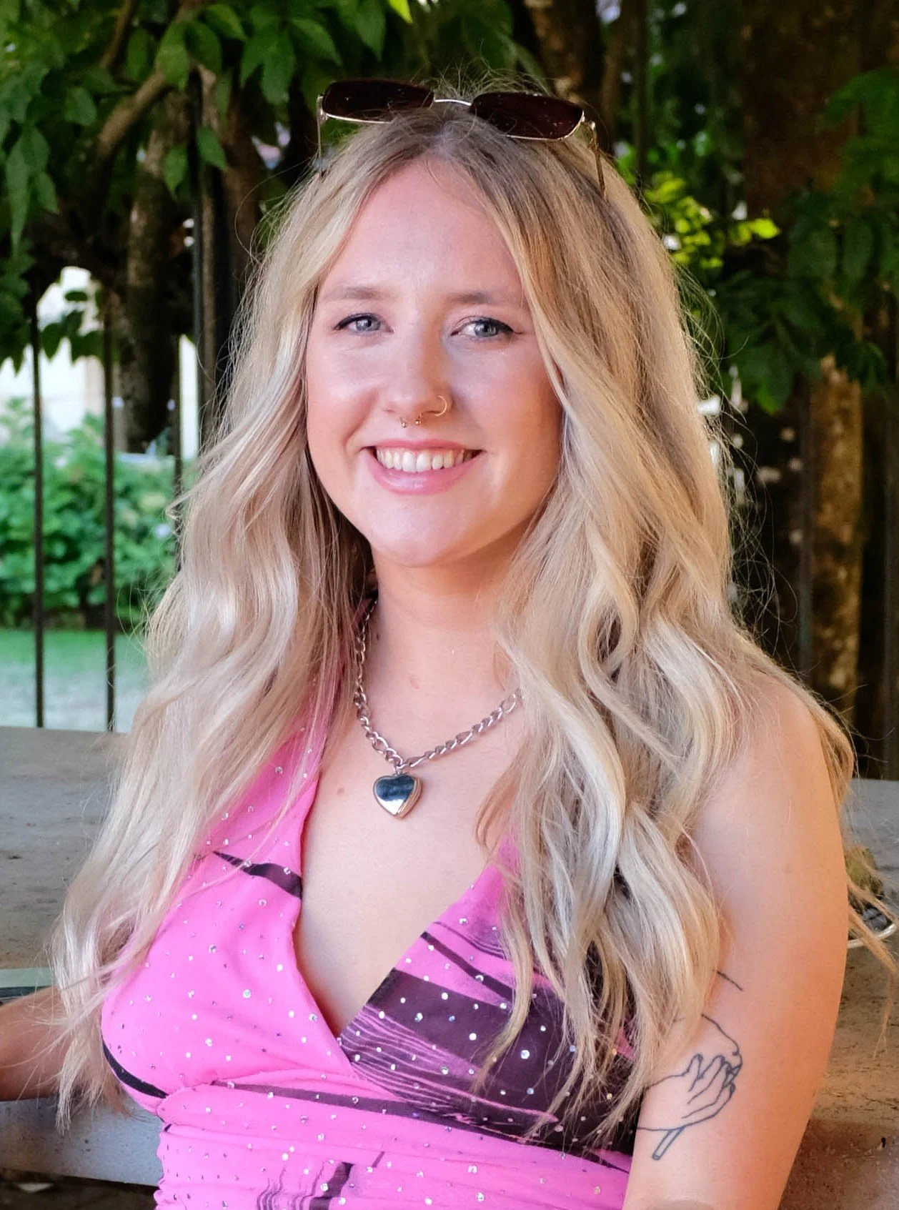 Young woman with long blonde wavy hair, wearing sunglasses on her head, a silver heart necklace, a pink polka dot dress, and a nose ring, smiling outdoors with greenery in the background.
