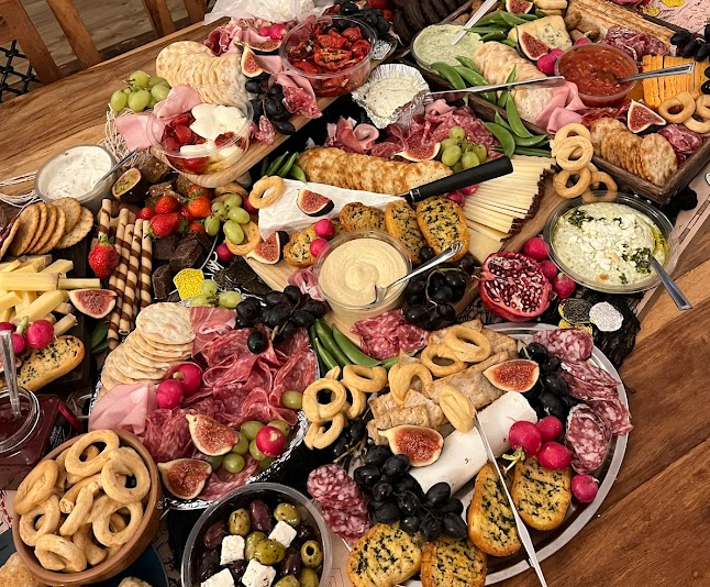A large spread of assorted cheeses, cured meats, fresh fruits like grapes and figs, crackers, bread, and various dips and spreads on a wooden table for a charcuterie board.