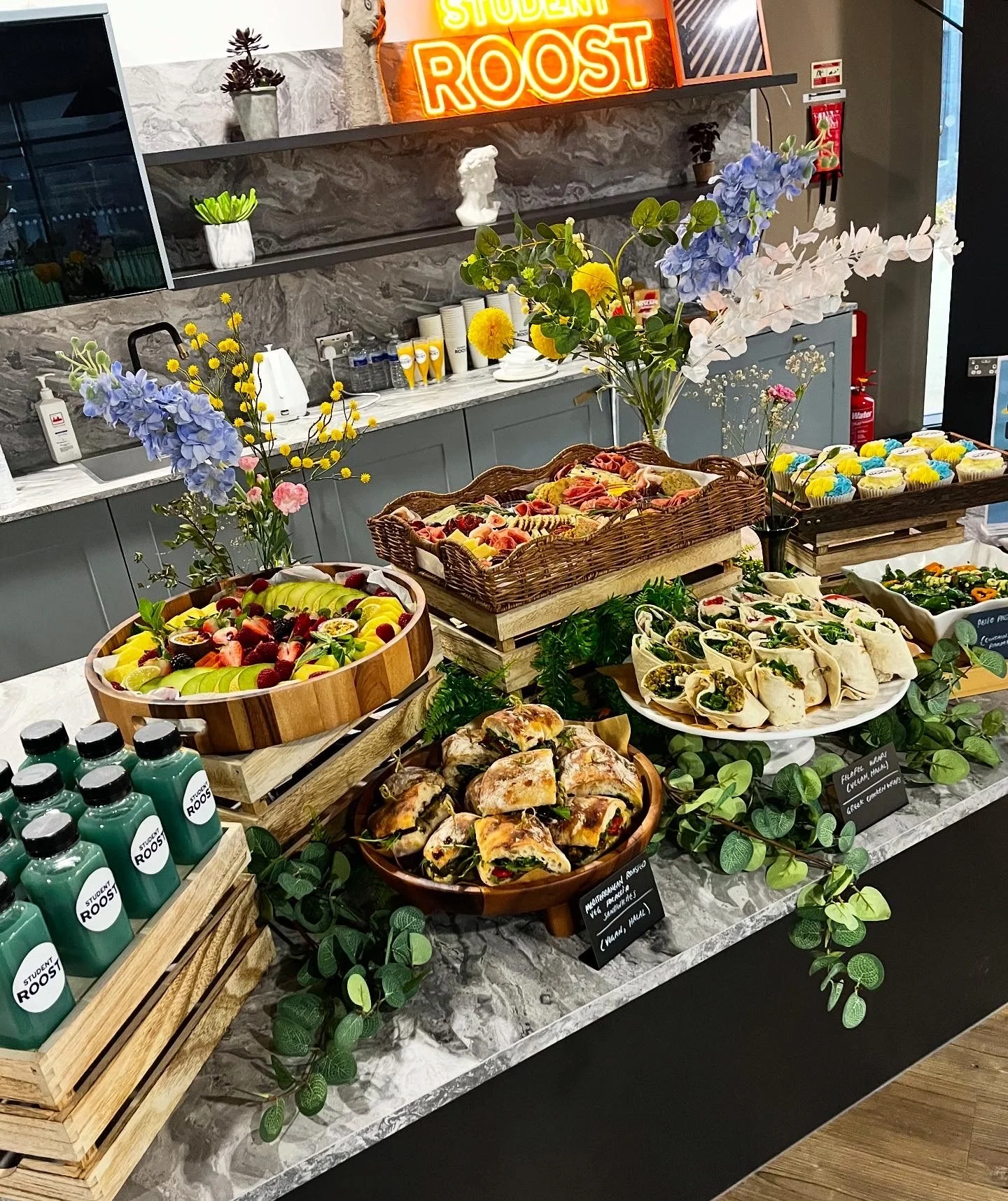 A long table with a black tablecloth displays an assortment of cheeses, cured meats, grapes, crackers, and dips, arranged for a buffet or tasting event. A sign on the table thanks partners such as Juicy Marbles and others.