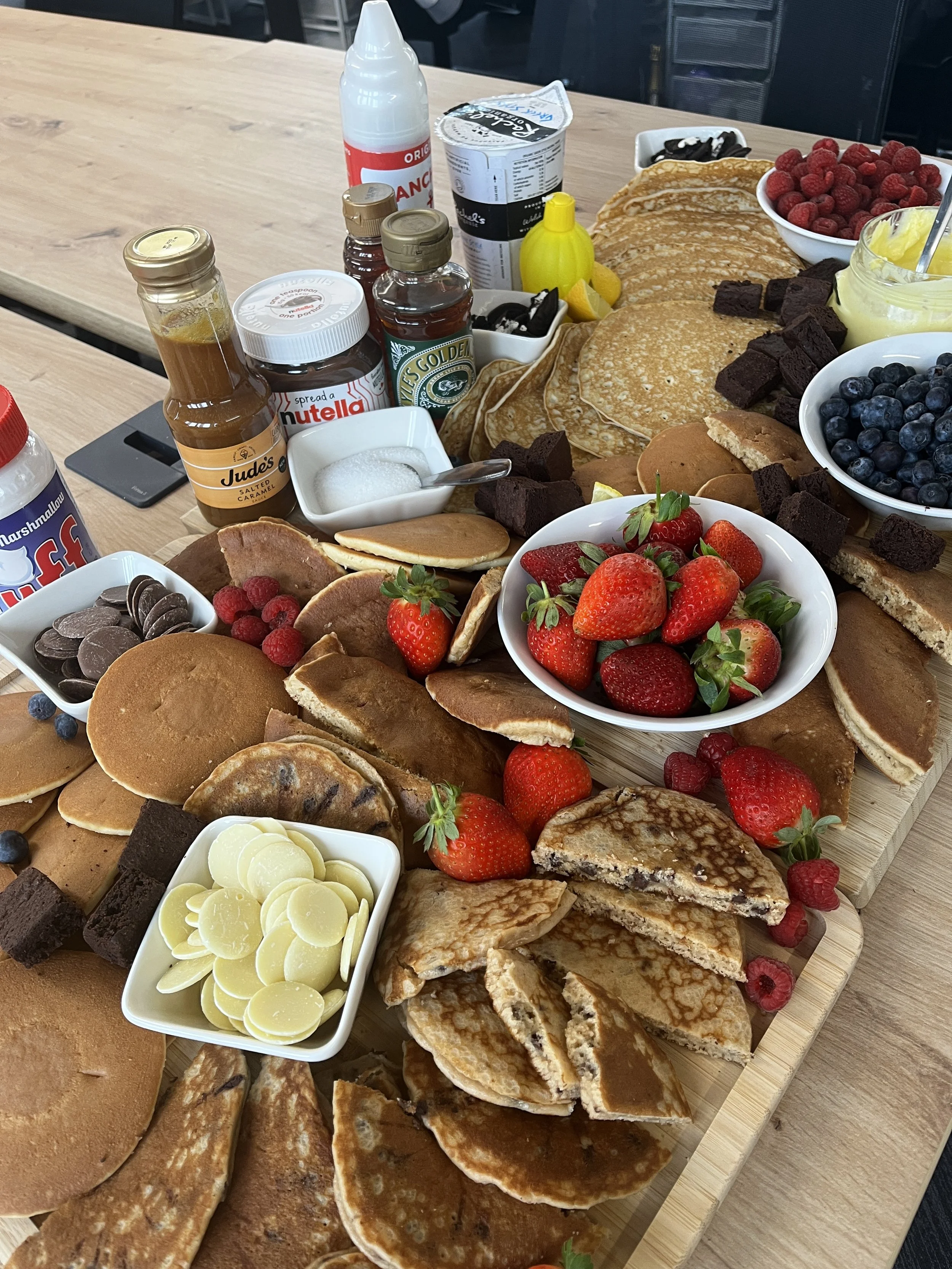 A large breakfast spread on a wooden table with pancakes, strawberries, blueberries, raspberries, chocolate pieces, syrup, Nutella, caramel sauce, whipped cream, and various condiments.