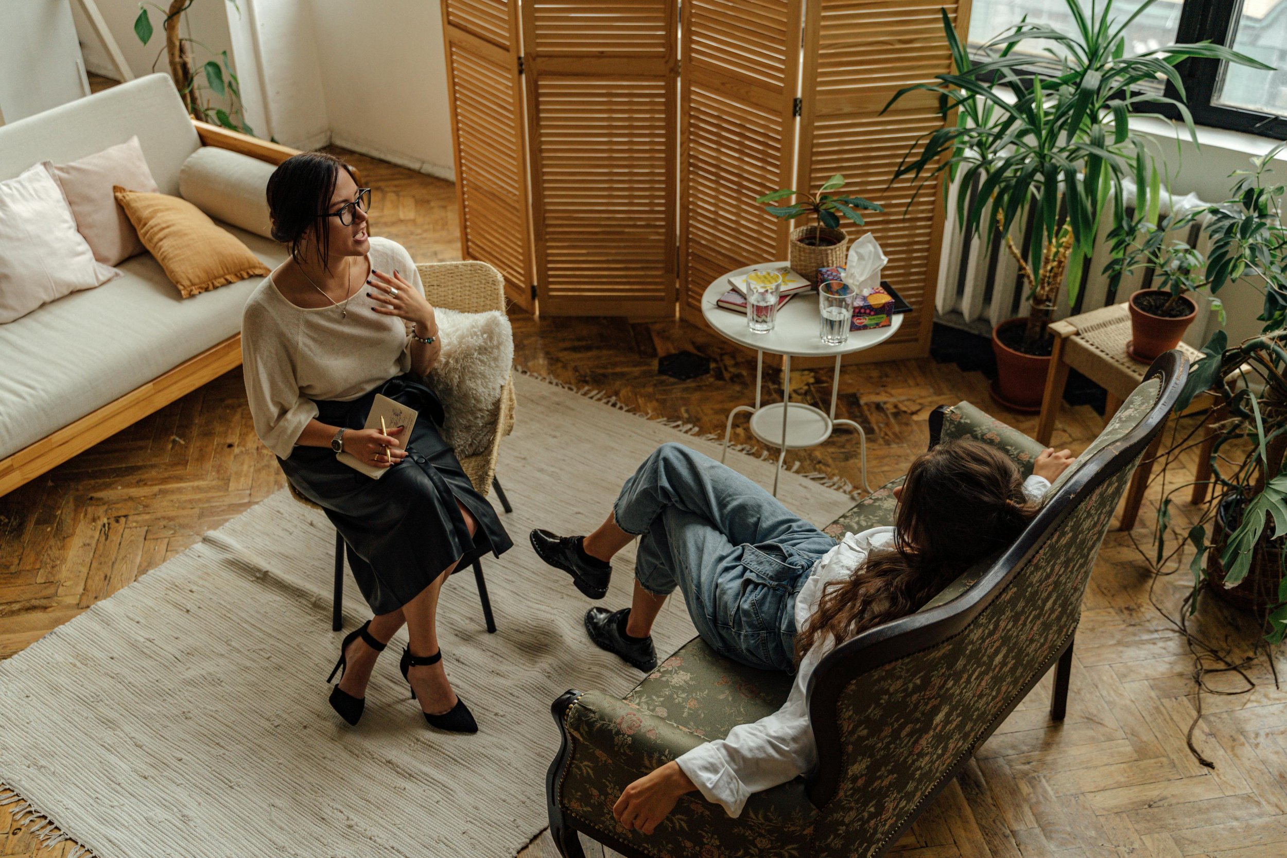 Two women having a conversation in a cozy living room with wooden flooring, a white sofa with beige and brown cushions, and a window with potted plants. One woman sits on a chair with a notebook and pen, the other reclines on a patterned sofa.