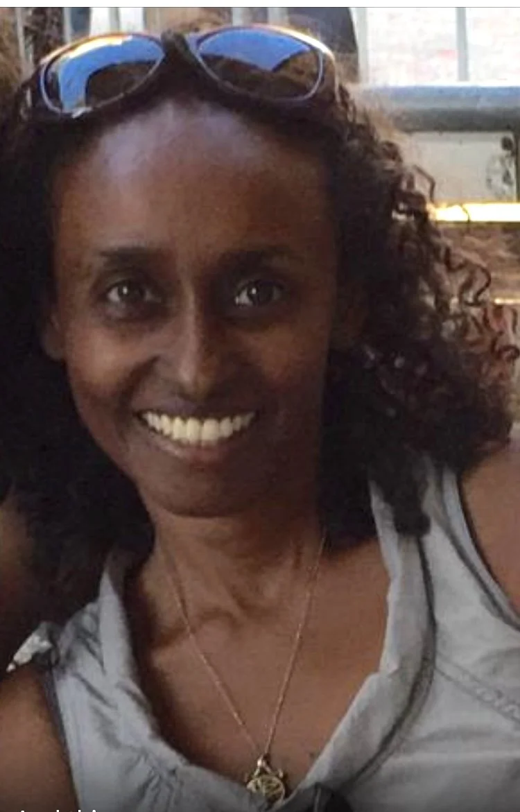 A smiling woman wearing sunglasses on her head, a light-colored sleeveless top, and a necklace, sitting indoors near a window.