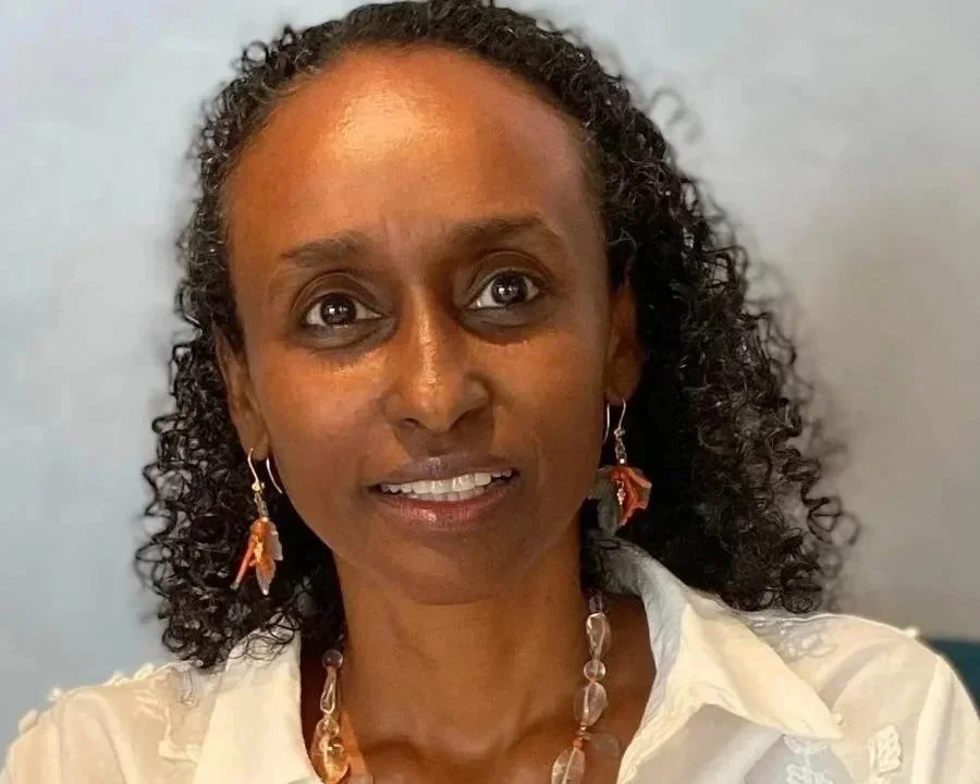 A woman with curly dark hair wearing earrings and a necklace, smiling at the camera.