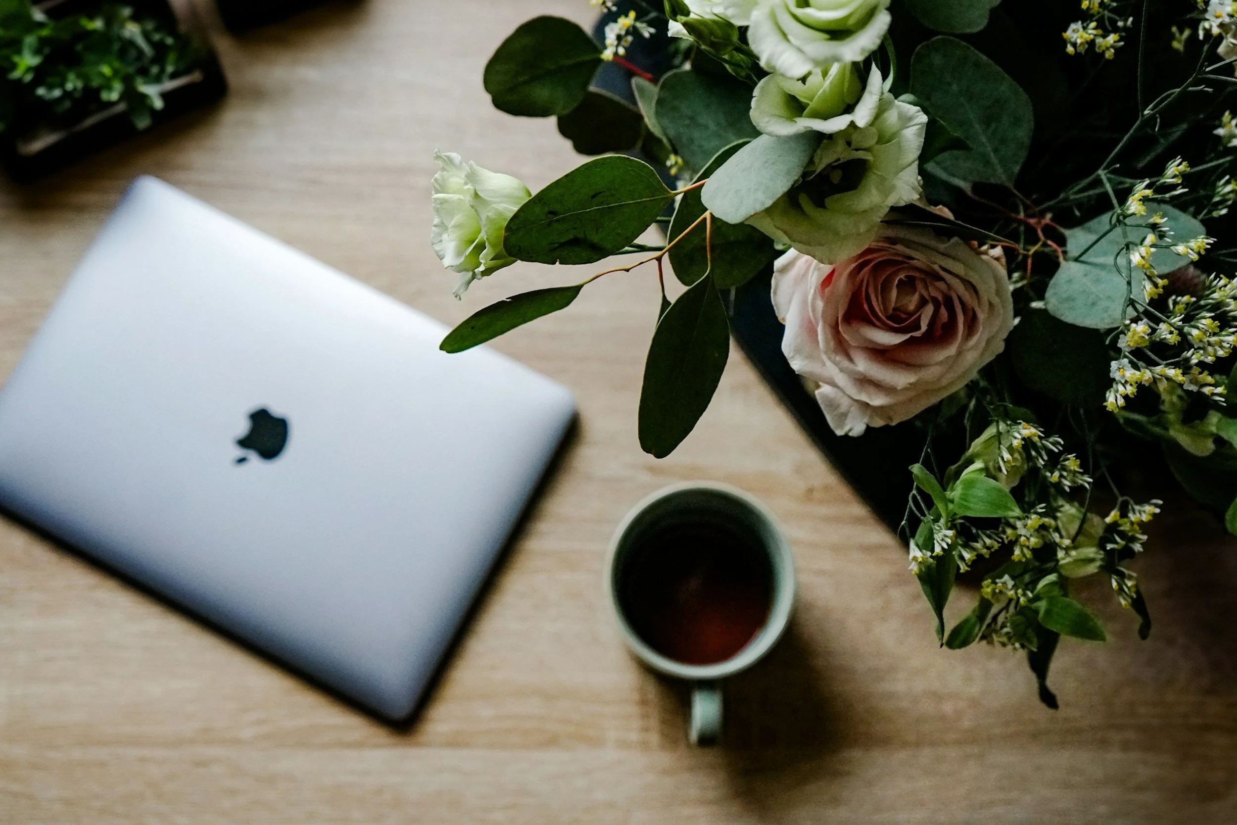 A top-down view of a wooden desk with a closed silver MacBook, a cup of coffee, and a large bouquet of flowers with roses and greenery.