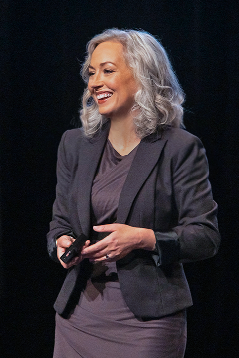A woman with gray, curly hair smiling while holding a remote or clicker, dressed in a dark blazer and dress, standing on a stage with a black background.