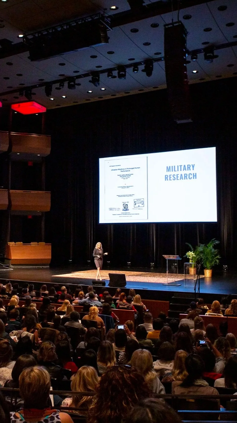 A woman giving a presentation on stage at a conference with a large audience seated and taking photos.