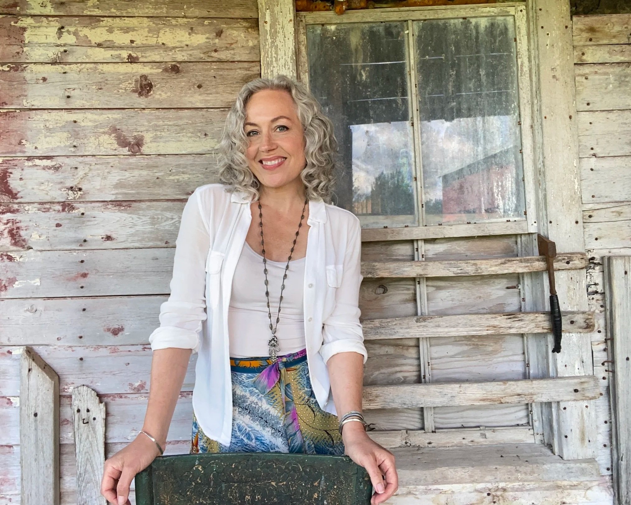 A smiling woman with curly gray hair wearing a white shirt, colorful skirt, and jewelry, standing in front of an old, weathered wooden wall with a window behind her.