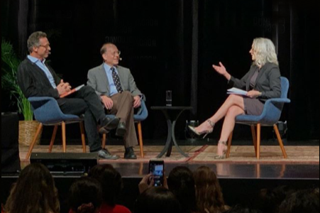 Three people seated on stage during a talk show or interview, with a woman holding papers and two men listening, in front of an audience.