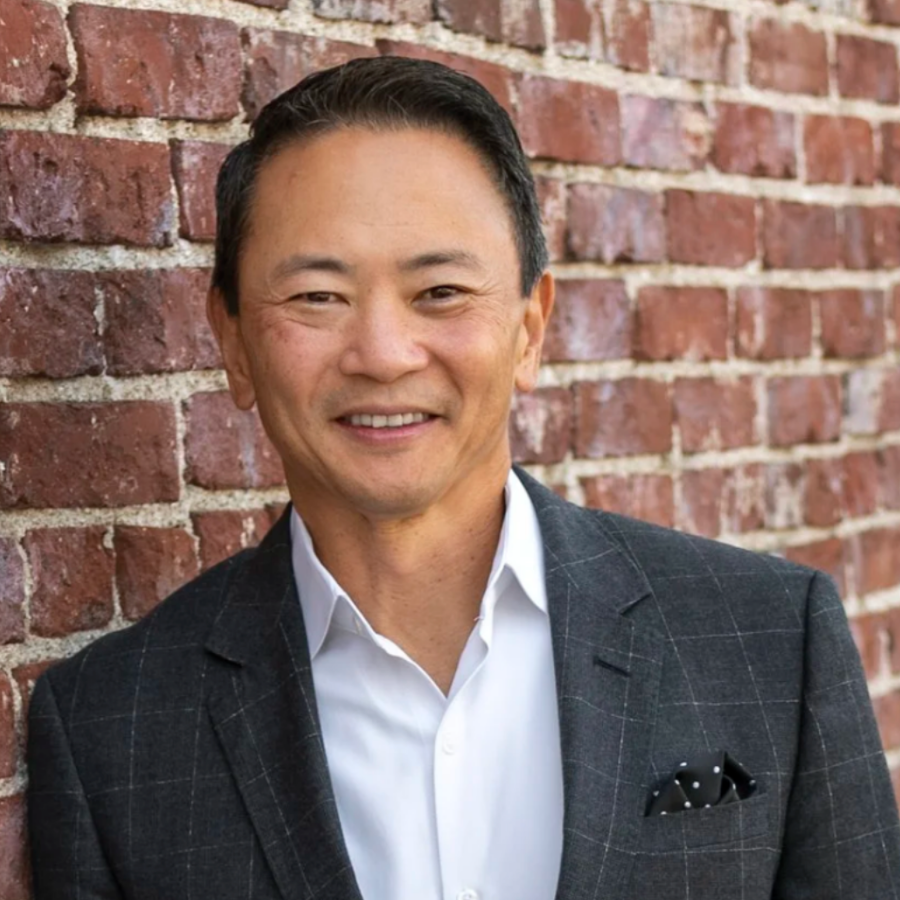 Portrait of a smiling Asian man in a white shirt and gray checkered blazer standing against a red brick wall.