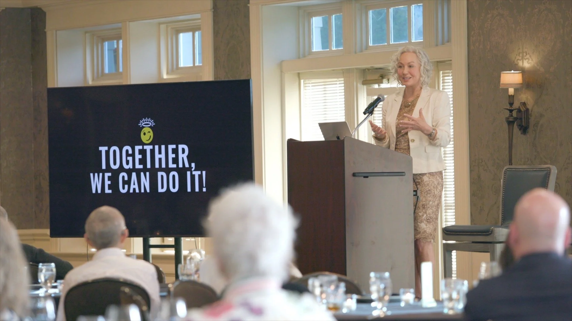 A woman with curly gray hair giving a presentation at a conference, standing behind a wooden podium with a laptop, in a room with large windows and vintage wallpaper. A large screen displays the message "Together, we can do it!" with a yellow smiley face and a halo above it. Audience members are seated at tables, some with glasses of water.