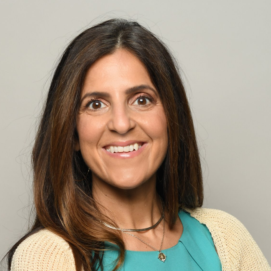 A smiling woman with long brown hair, wearing a teal top, cream cardigan, and layered necklaces, against a plain gray background.