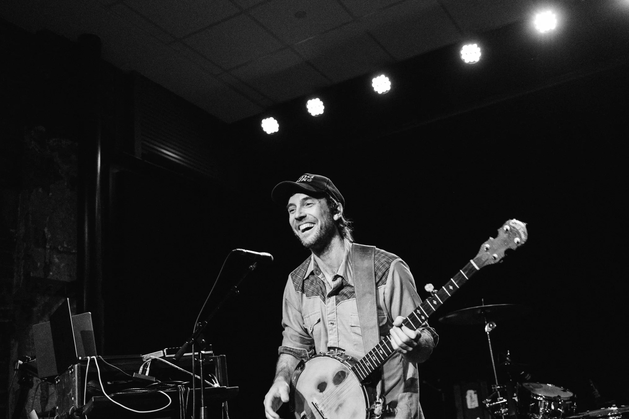 A man smiling and playing an acoustic guitar on a dimly lit stage, with stage lights overhead and musical equipment visible.