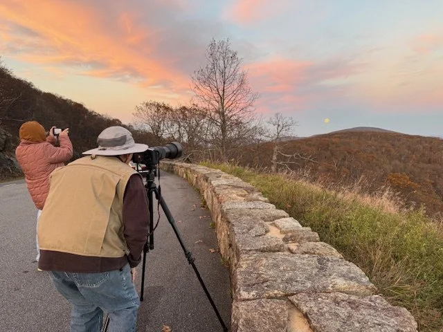 Two people standing on a mountain road during sunset, one person with a camera on a tripod looking at the horizon, and the other person taking a photograph with a phone, with colorful sky and bare trees in the background.