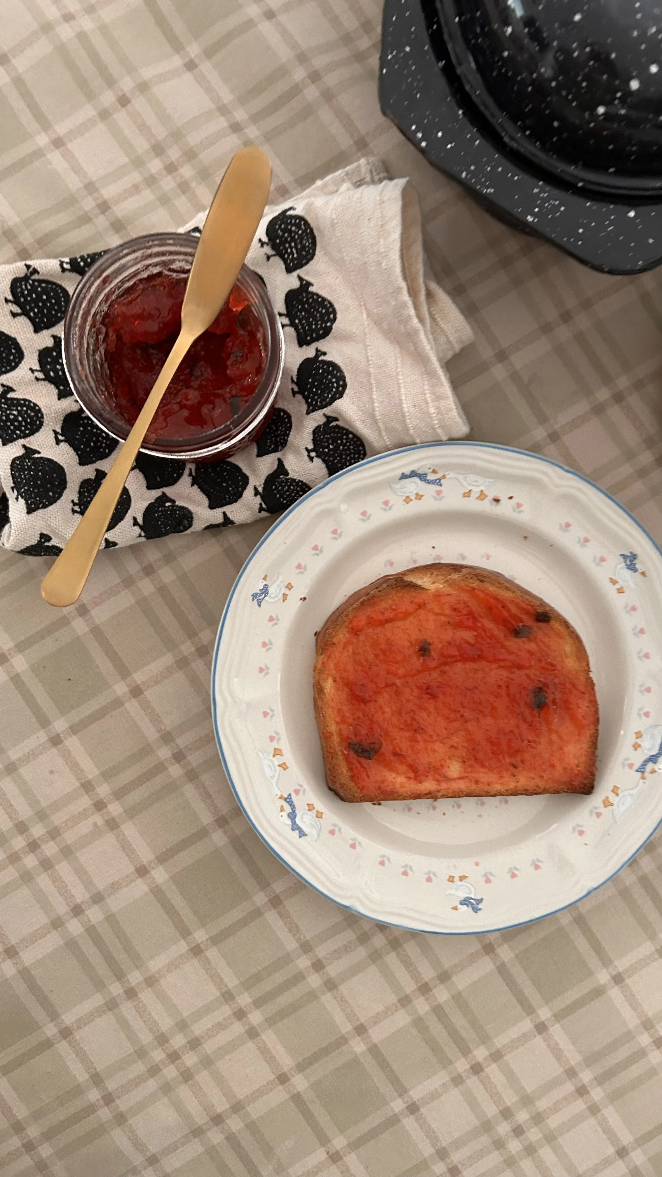 Slice of toast with tomato sauce on a white plate with blue trim, jar of strawberry jam with a gold knife, and a black speckled appliance on a checkered tablecloth.