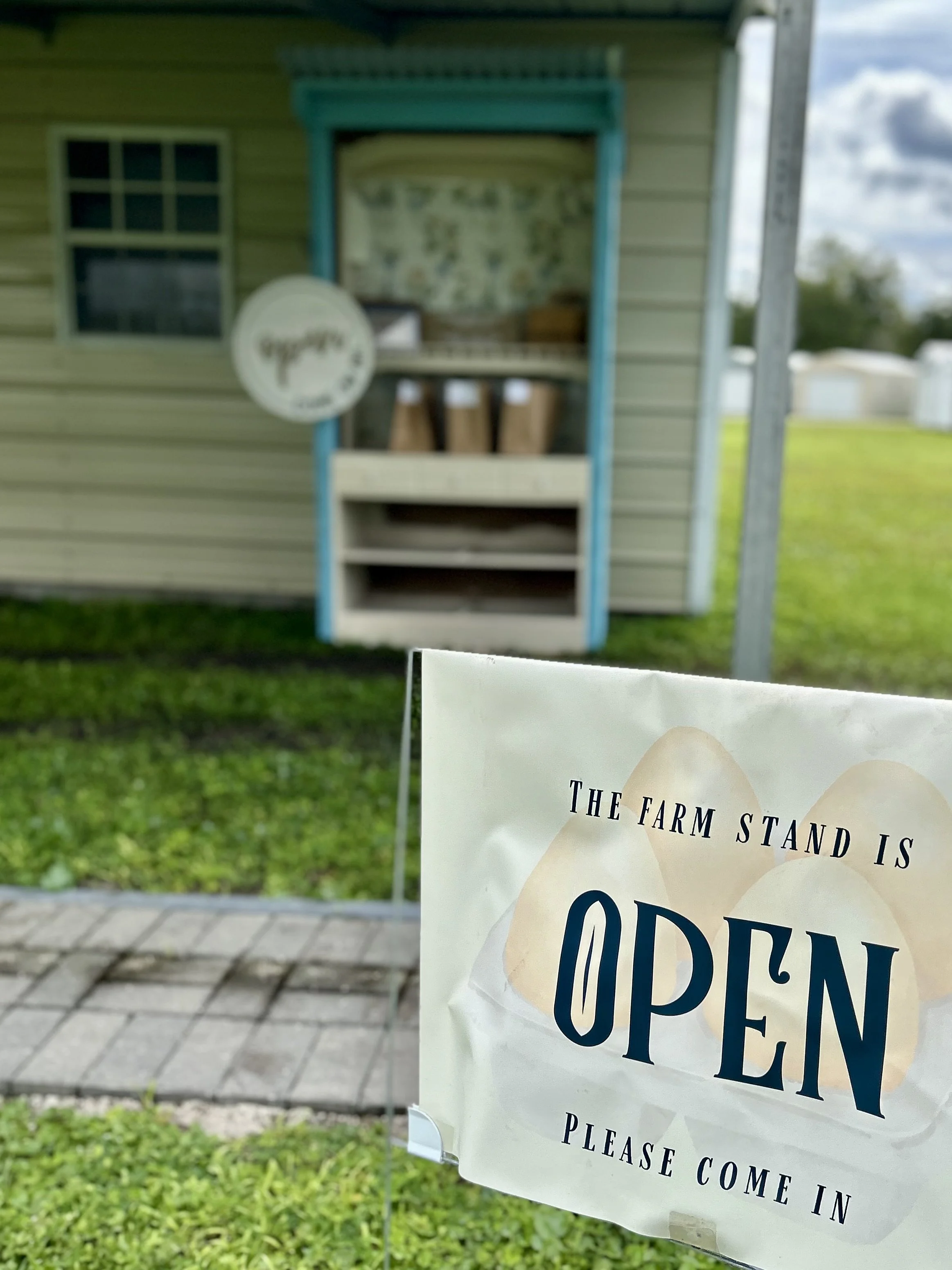Close-up of a sign that reads 'The farm stand is open, please come in,' with a farm stand in the background with some containers or jars.
