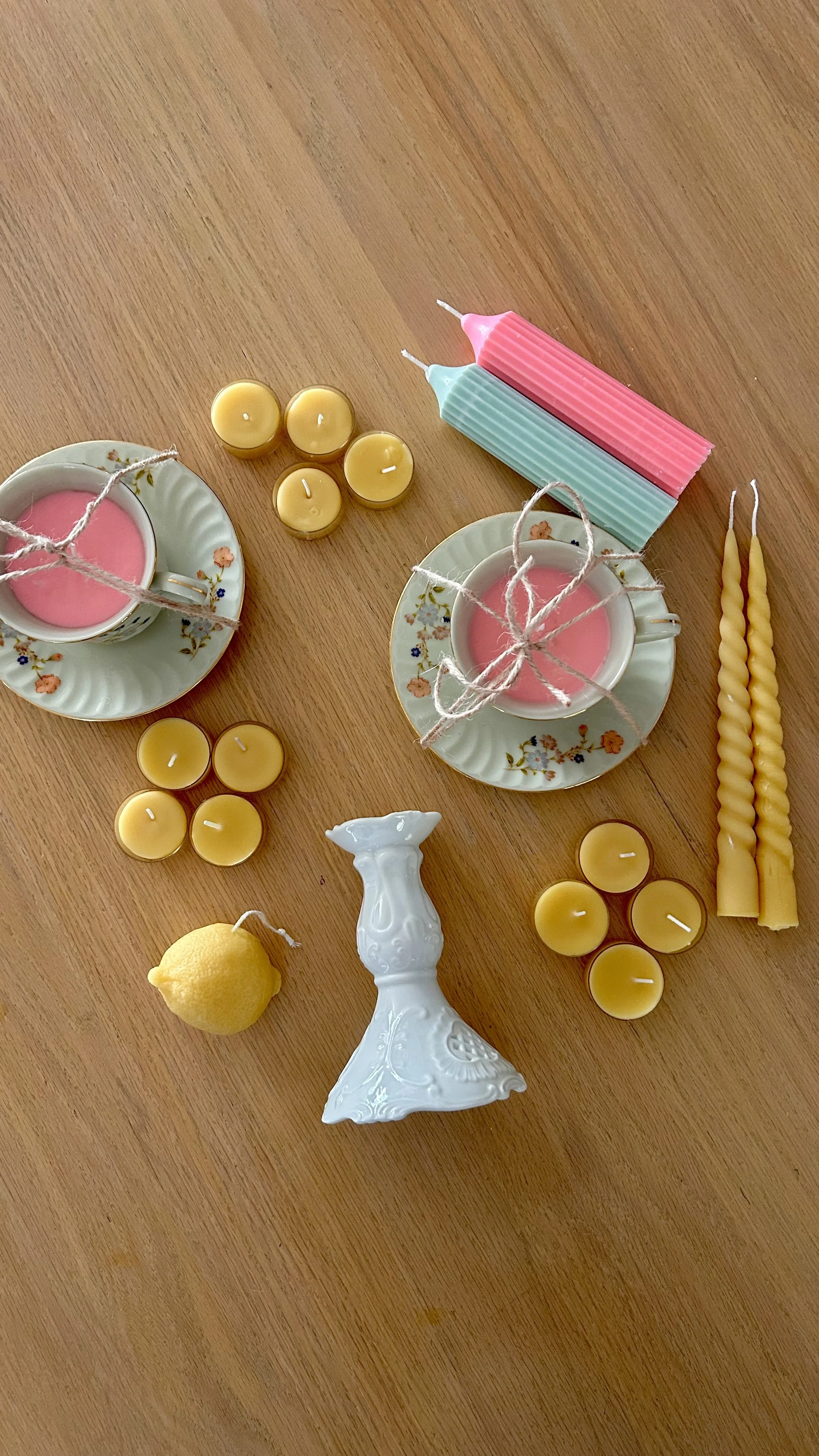 Decorative table arrangement with pink candles in teacups, yellow candles, twisted yellow candles, a white ceramic vase, a lemon, and pastel-colored candles on a wooden surface.