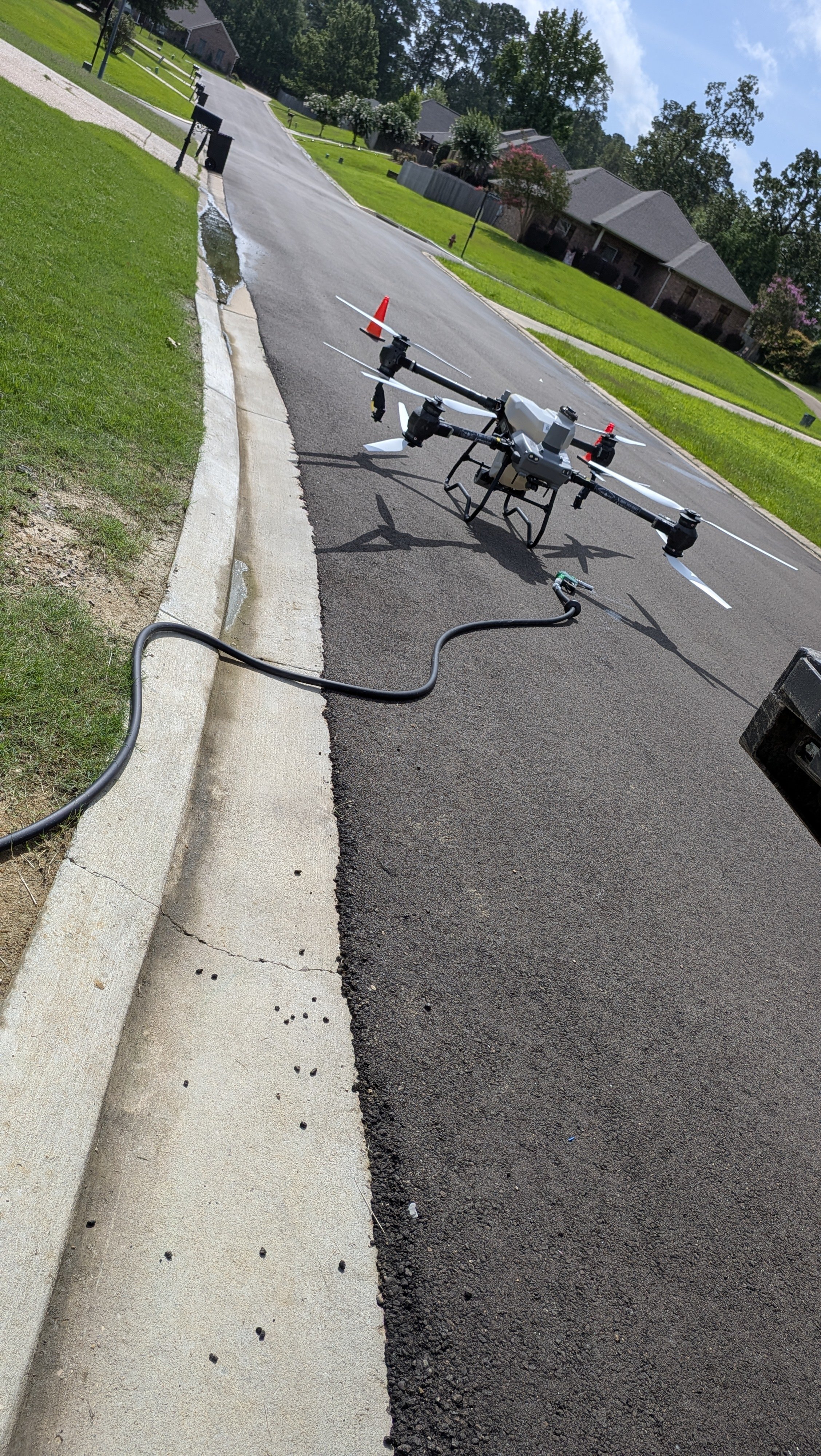 An unmanned drone on a paved street, connected to a power source or charging station, with a residential neighborhood in the background.