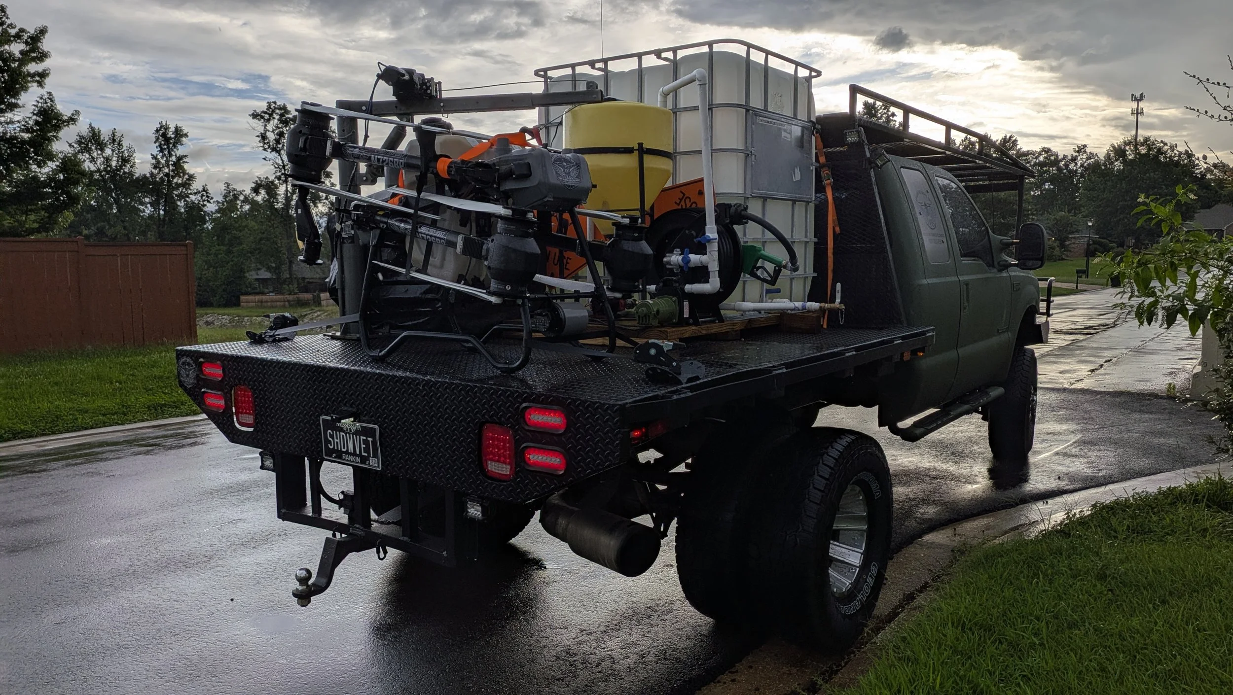 A green pickup truck equipped with water spraying equipment on the back, parked on a wet road during cloudy weather.