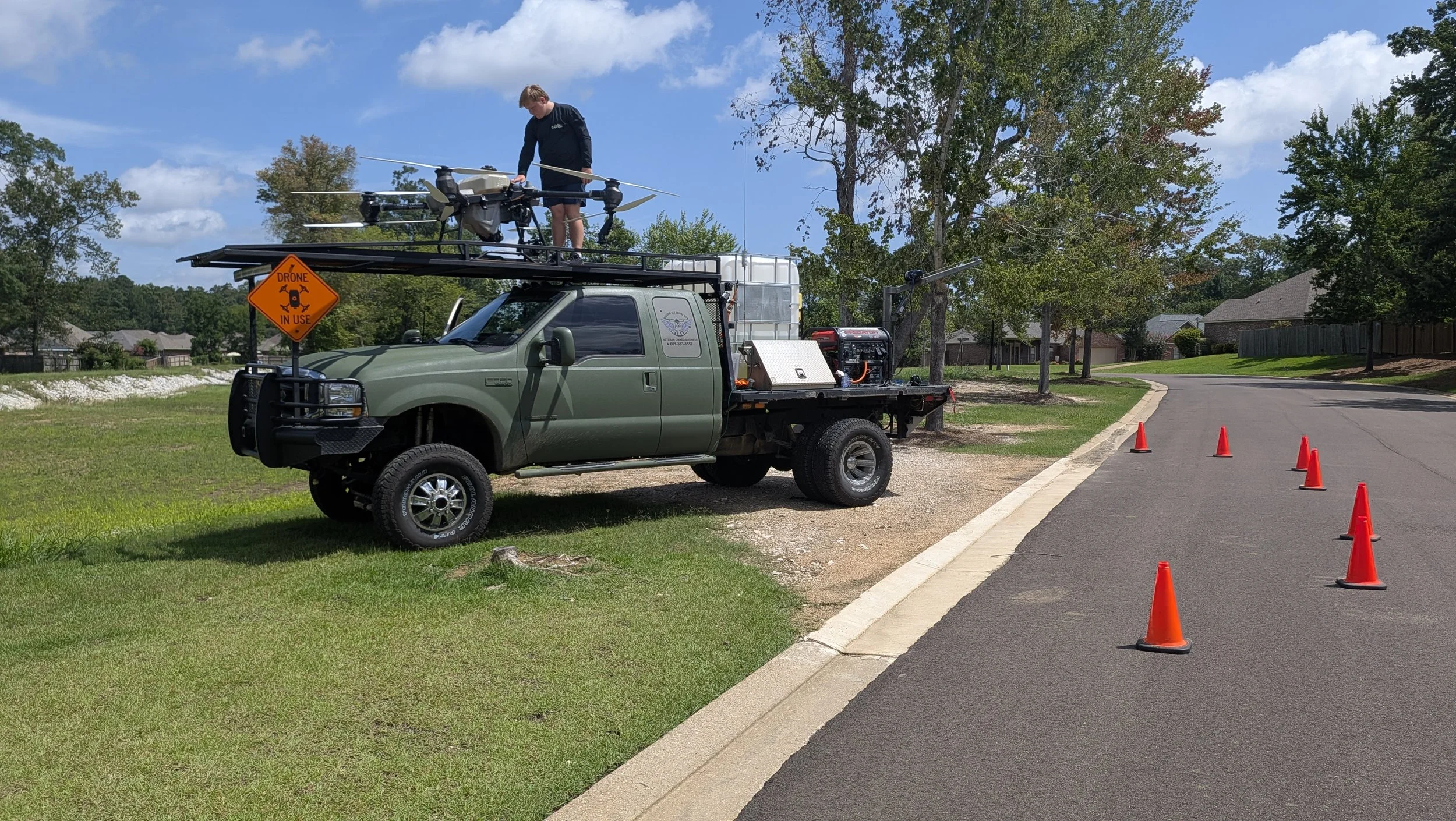 A green utility truck equipped with drone technology on a suburban street with orange cones and a signal line, man on top of the truck with drone equipment, residential houses and trees in the background.