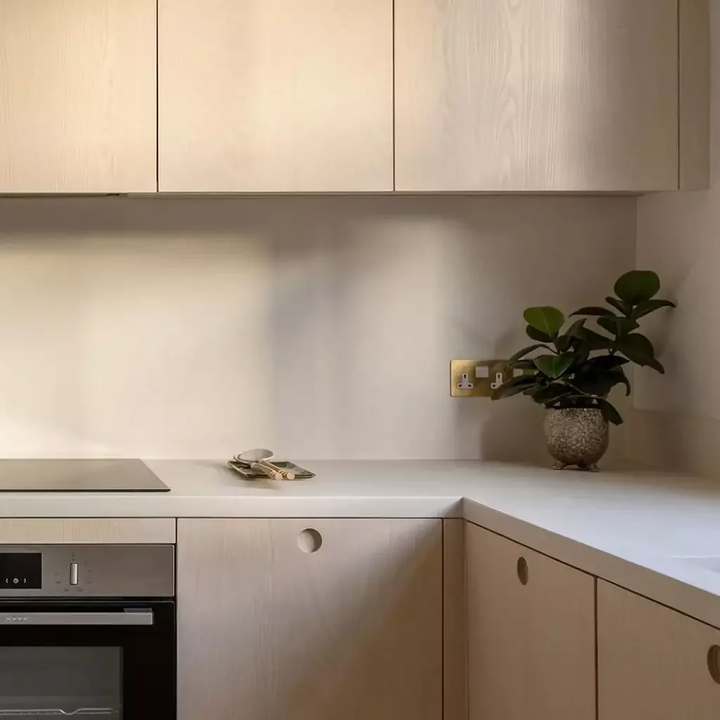 A minimalist kitchen with light wood cabinets, a white countertop, a small potted plant, and folded cloths on the counter.