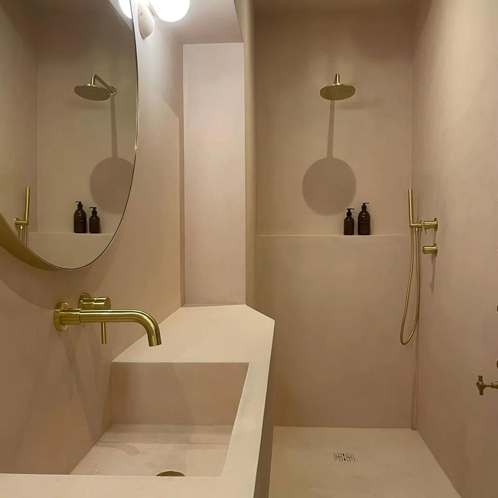 Minimalist beige bathroom with a large round mirror, gold fixtures, a built-in sink, and a separate shower area with a rain showerhead and handheld shower, two brown soap dispensers on a shelf.