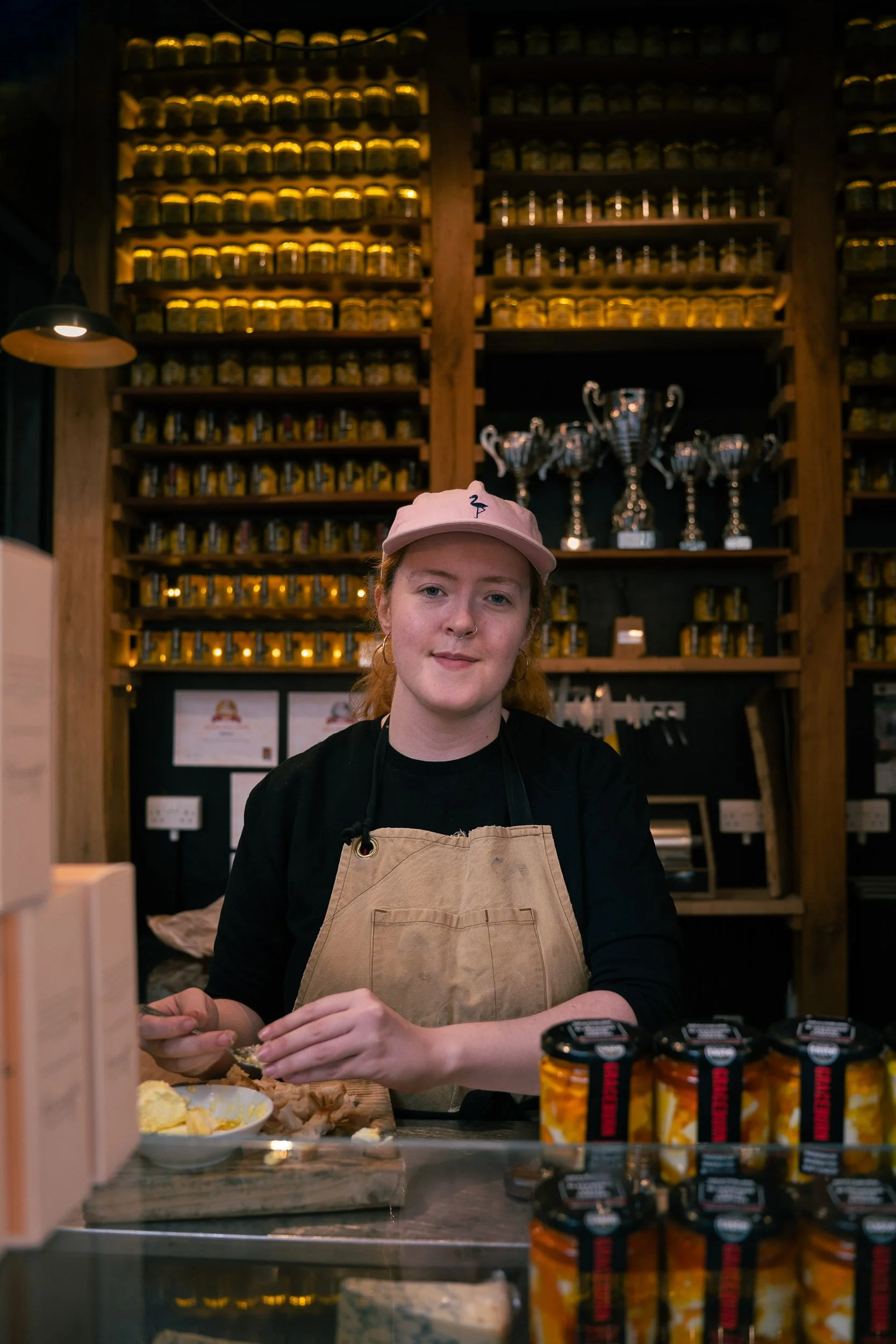 A woman with red hair, wearing a pink cap and a beige apron, standing behind a counter in a shop or cafe, surrounded by shelves filled with jars and trophies in the background.